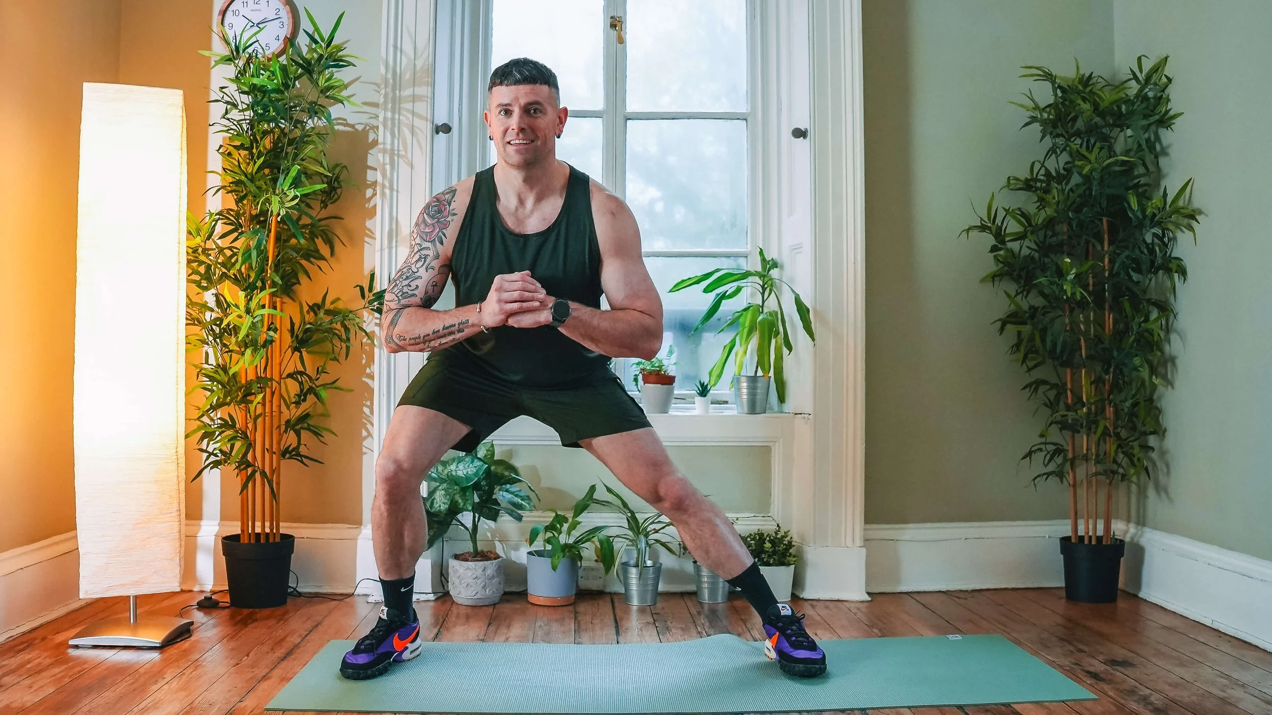 A man in athletic clothing performing a wide squat exercise on a yoga mat in a room with houseplants, a window, and a standing lamp.