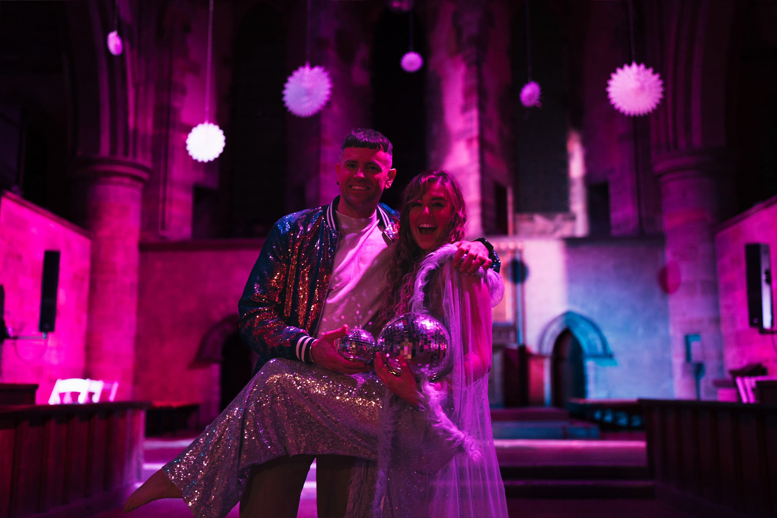 Two people celebrating at a party in a dimly lit room with pink and purple lighting, holding disco balls, with hanging decorations in the background.