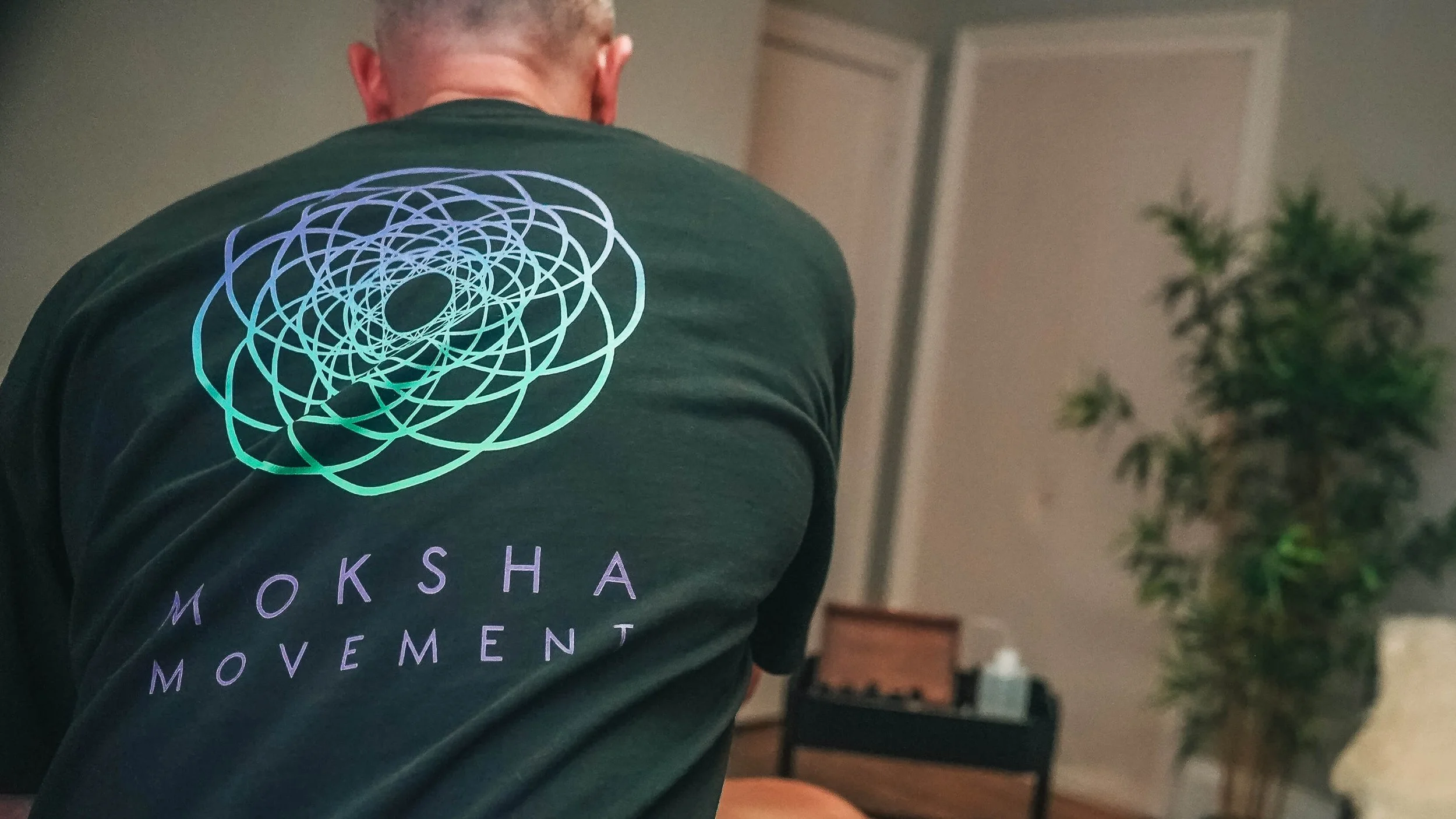Back of a person wearing a dark T-shirt with a colorful geometric design and the text 'MOKSHA MOVEMENT' printed on it, indoors with a blurred background of a plant and furniture.