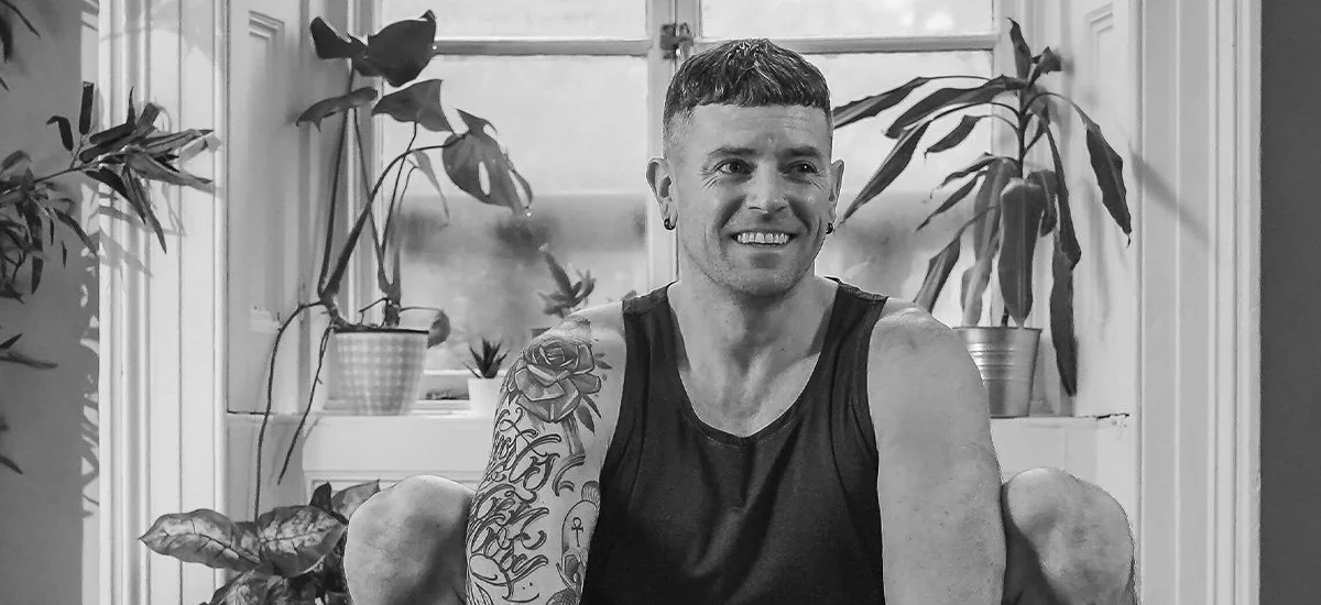 A smiling man with tattoos on his right arm, wearing a sleeveless black shirt, sitting inside a room with potted plants by a window.