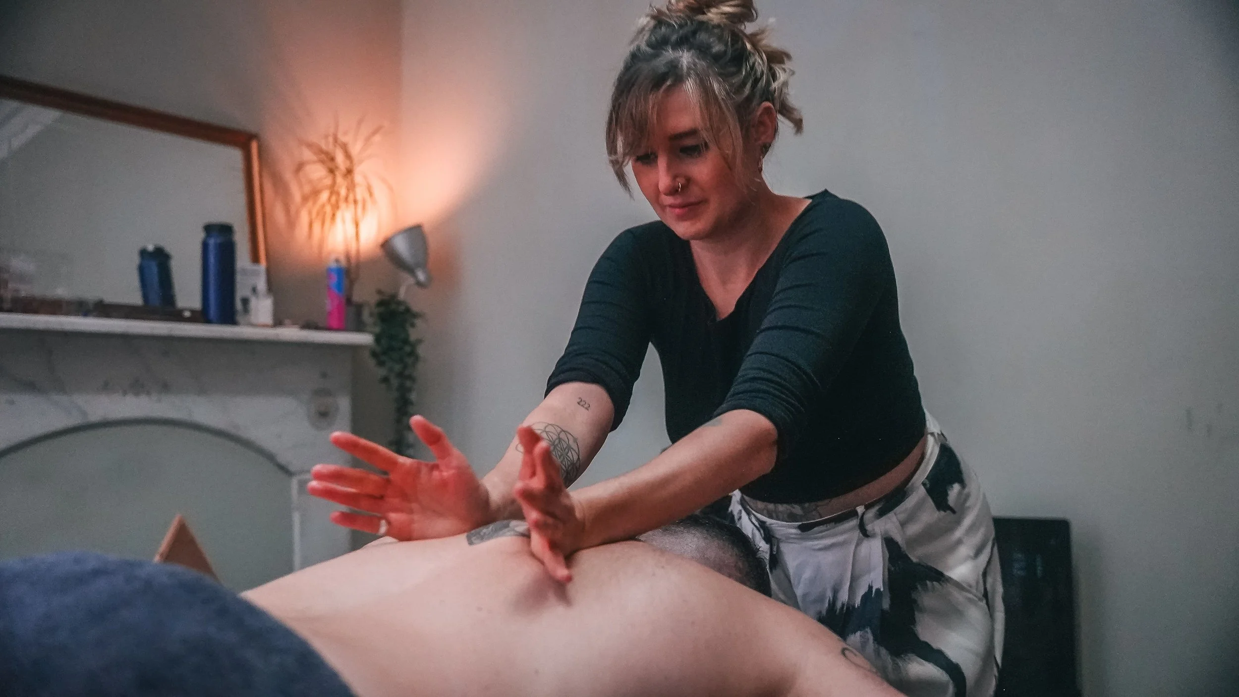A woman with short hair and tattoos on her arm is giving a massage to a person lying face down.