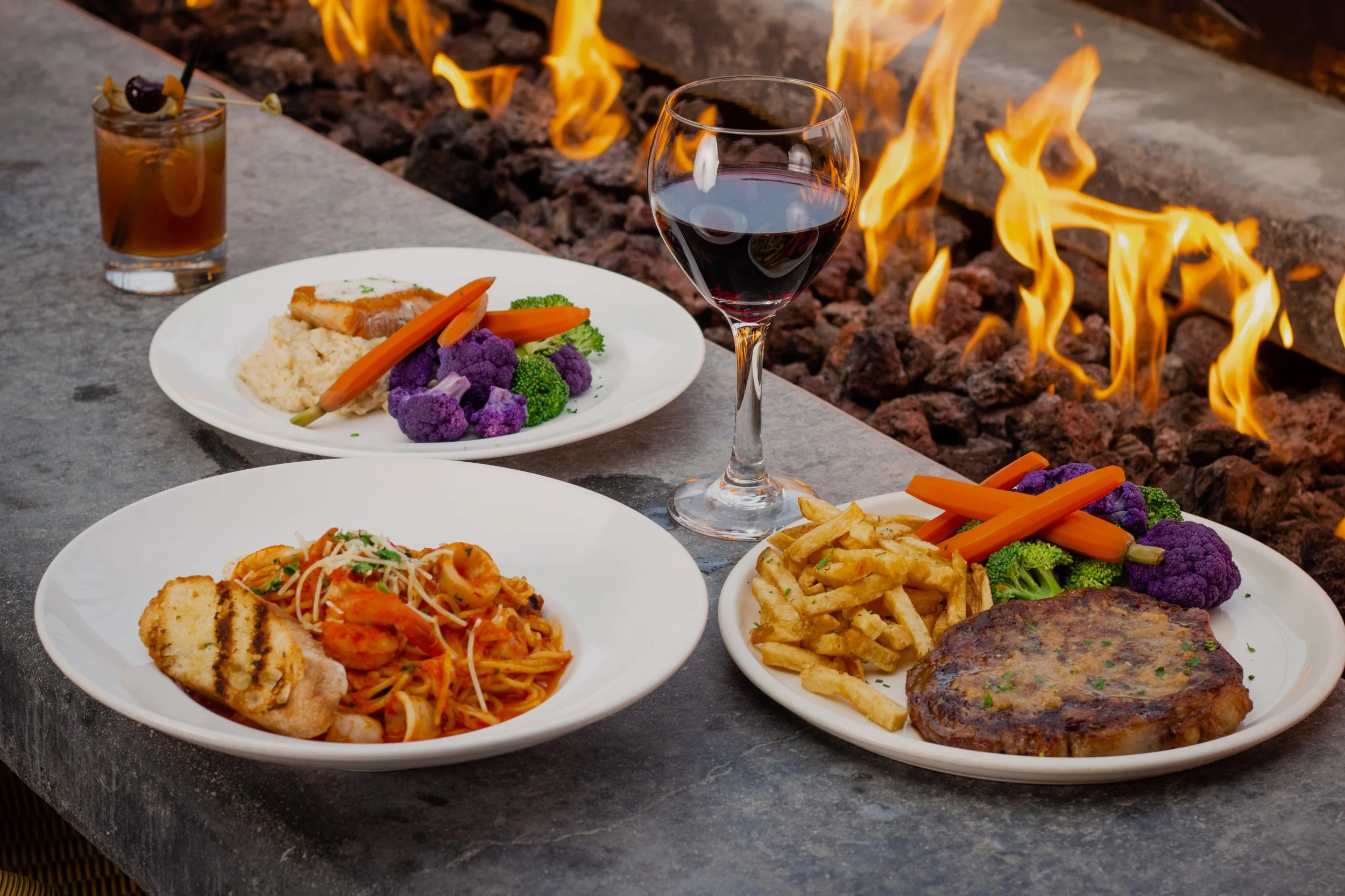A meal outdoors with a fire pit, featuring plates of pasta, a grilled steak with fries, a glass of red wine, and a cocktail, with flames in the background.