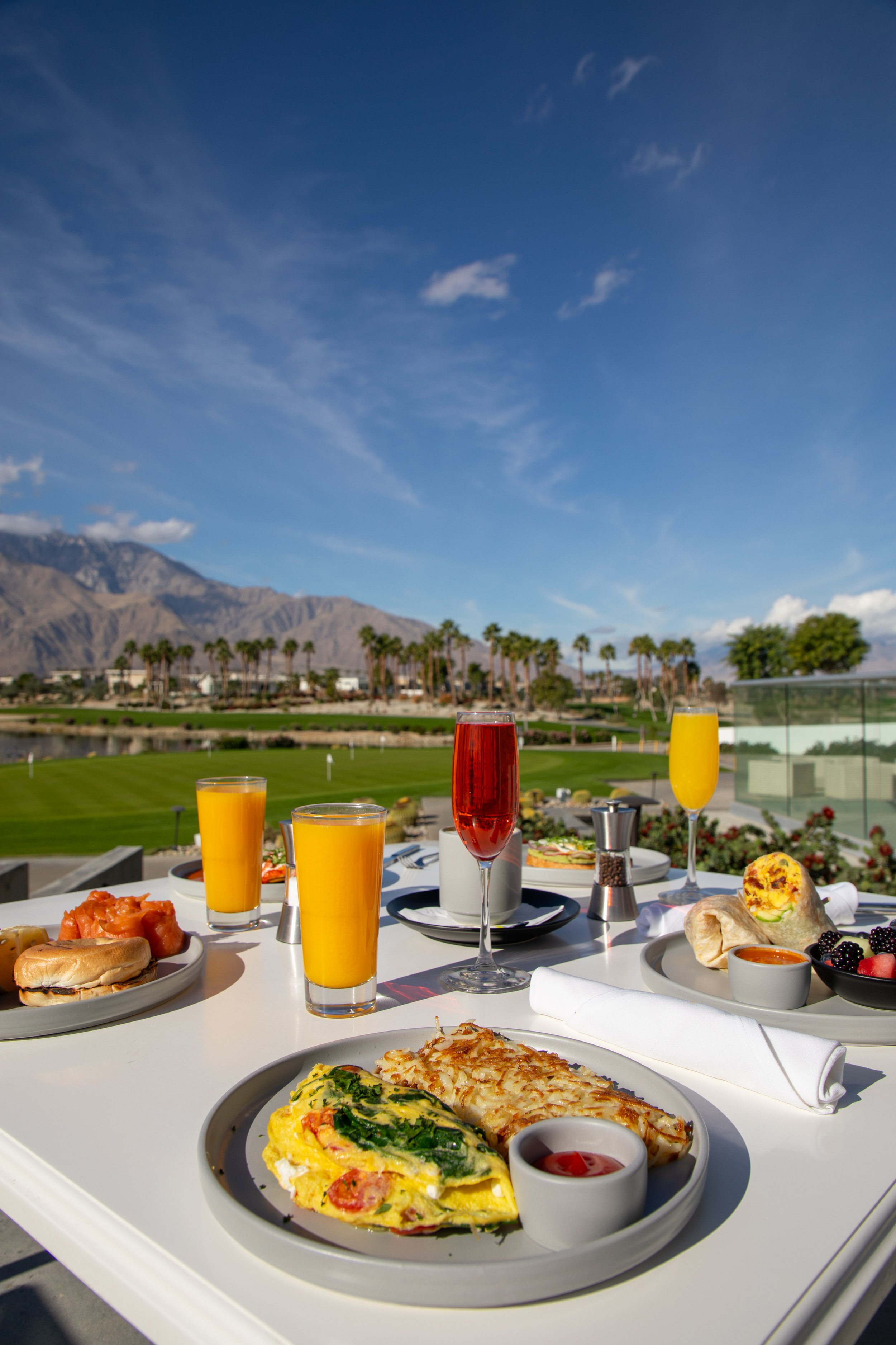 A luxurious outdoor breakfast table with plates of eggs, pastries, fruit, and beverages, set against a scenic backdrop of palm trees, mountains, and a blue sky with some clouds.