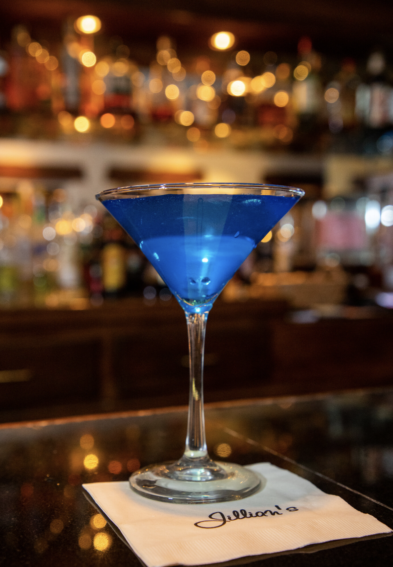 A blue-colored cocktail in a martini glass placed on a white napkin with the word 'Jillian's' written on it, inside a bar with warm, blurry, glowing lights in the background.