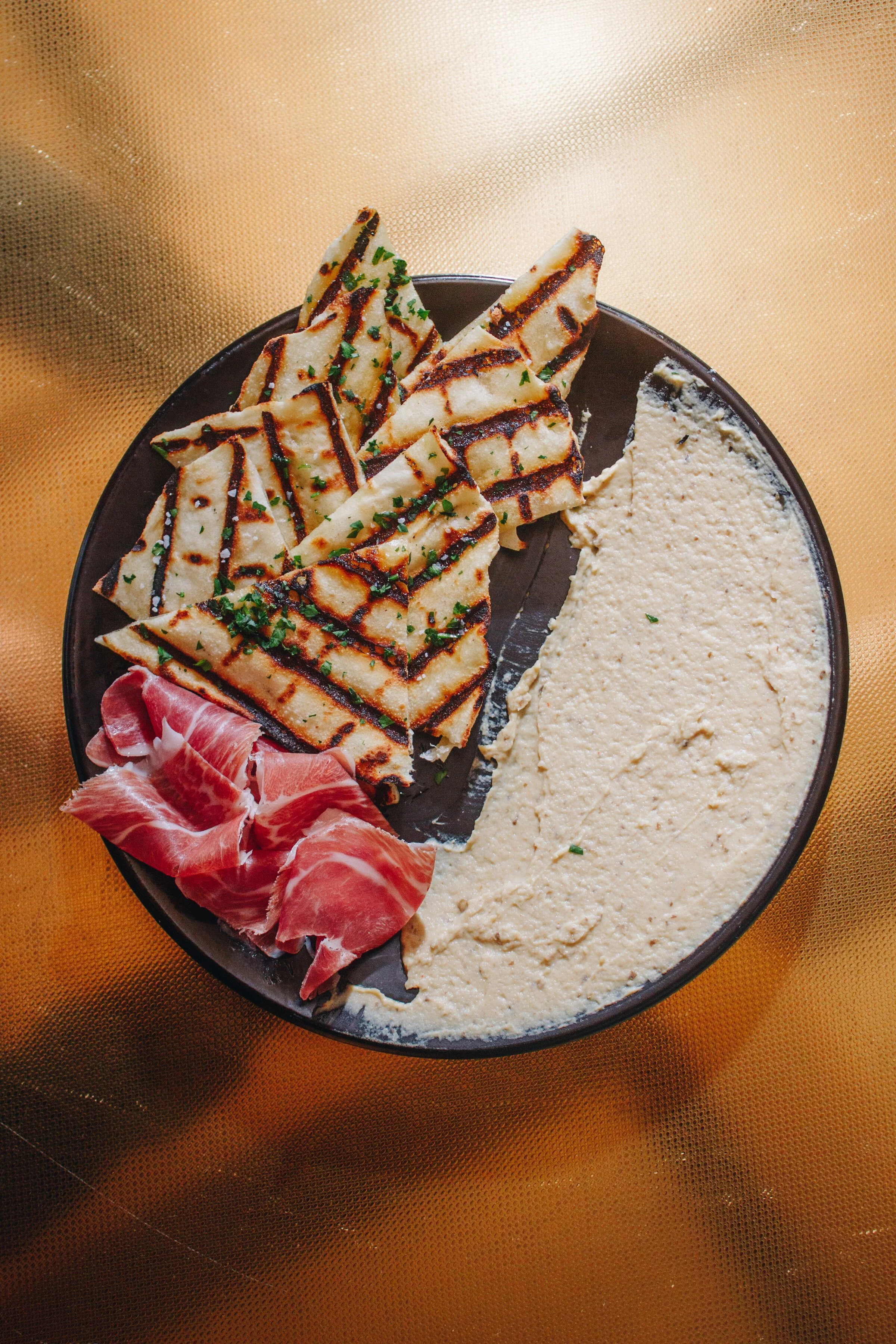 A black plate with grilled flatbread, a creamy white dip, and slices of prosciutto on a gold tablecloth.