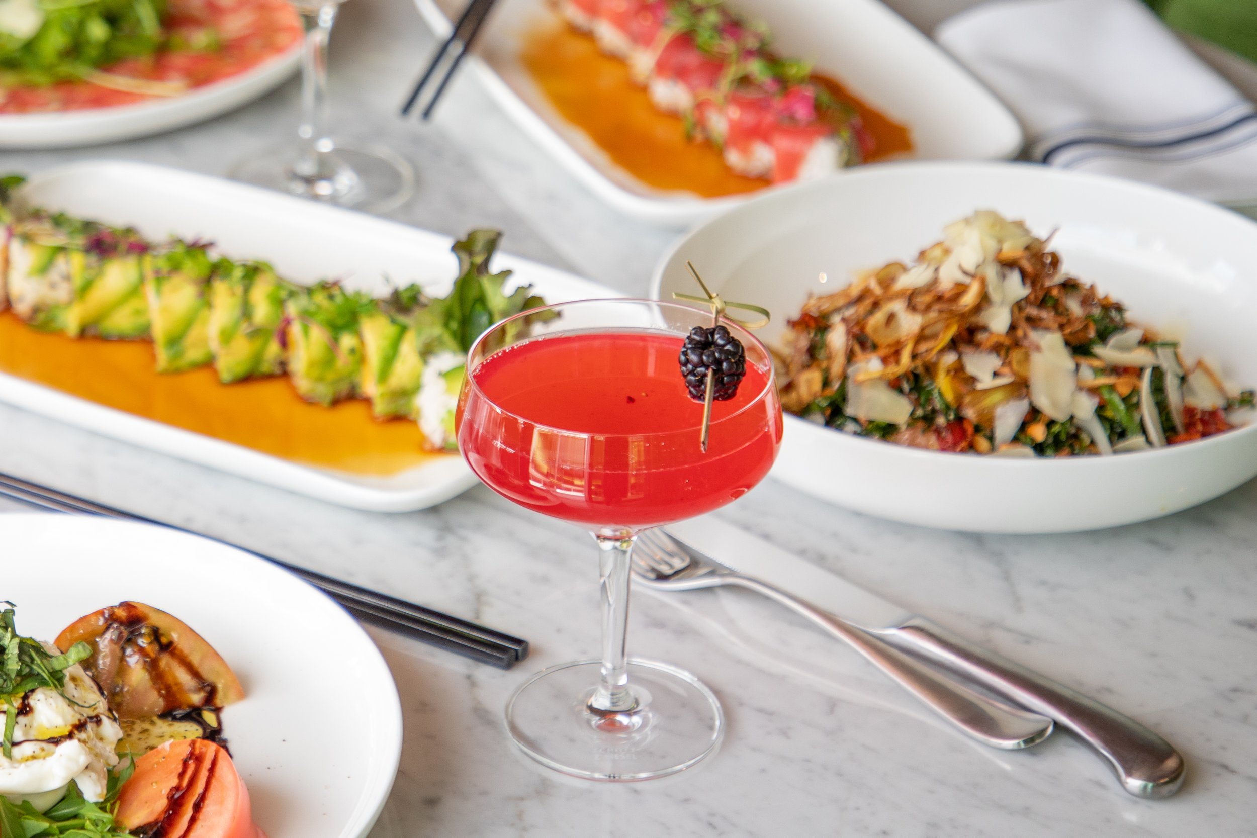 A table with various dishes, including a red cocktail with a blackberry garnish, sushi rolls on a white platter, a bowl of salad with shredded vegetables and cheese, and another sushi or appetizer dish in the background.