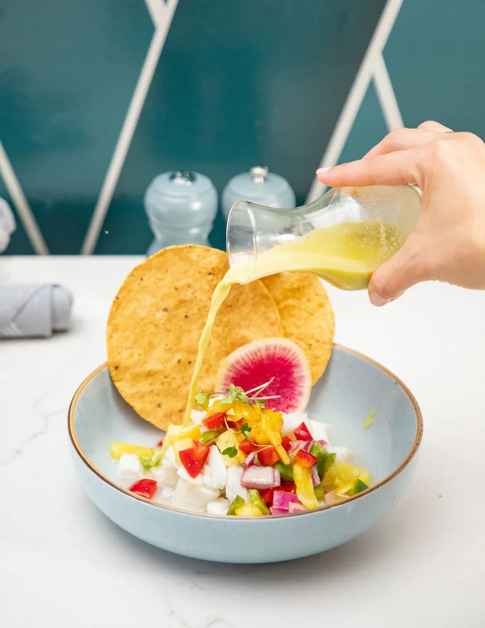 Watermelon radish, colorful chopped vegetables, and cheese in a bowl with nacho chips, as yellow dressing is poured over the salad.