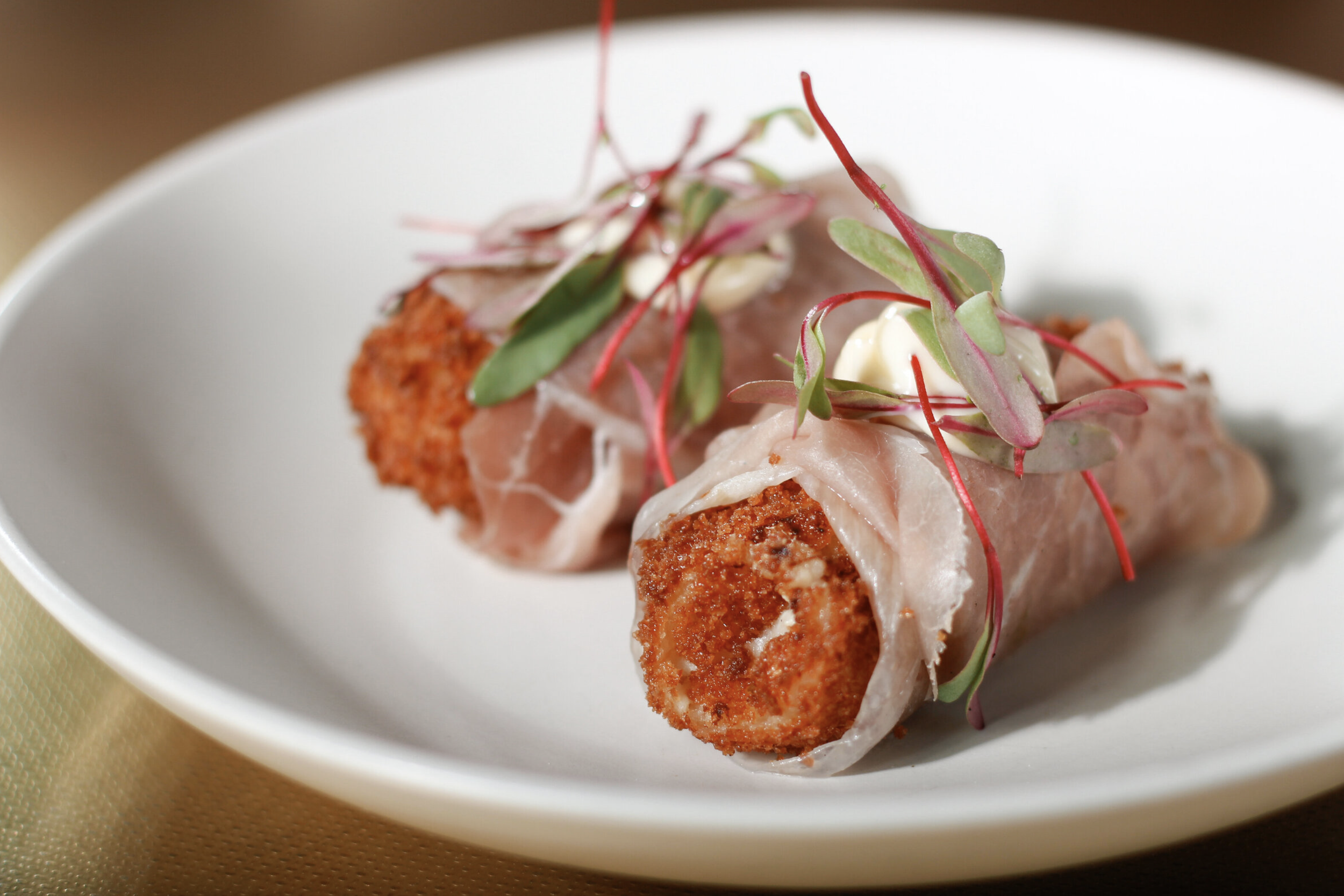 Two croquettes wrapped in prosciutto, topped with microgreens and a dollop of mayonnaise on a white oval plate.