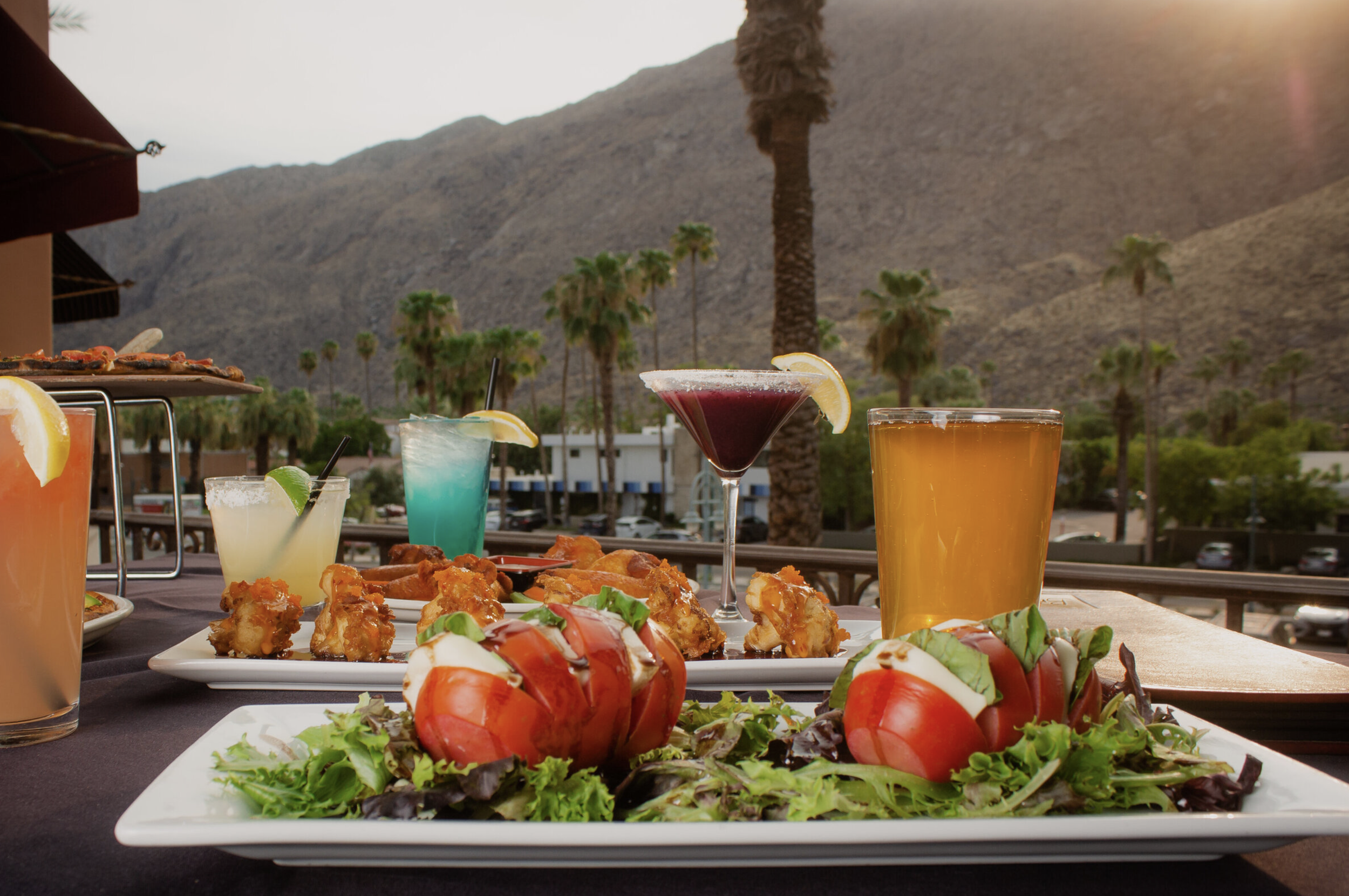 Outdoor dining table with various drinks and food, including a tomato and mozzarella salad, fried chicken, and colorful cocktails, with a mountainous desert landscape and palm trees in the background during sunset.