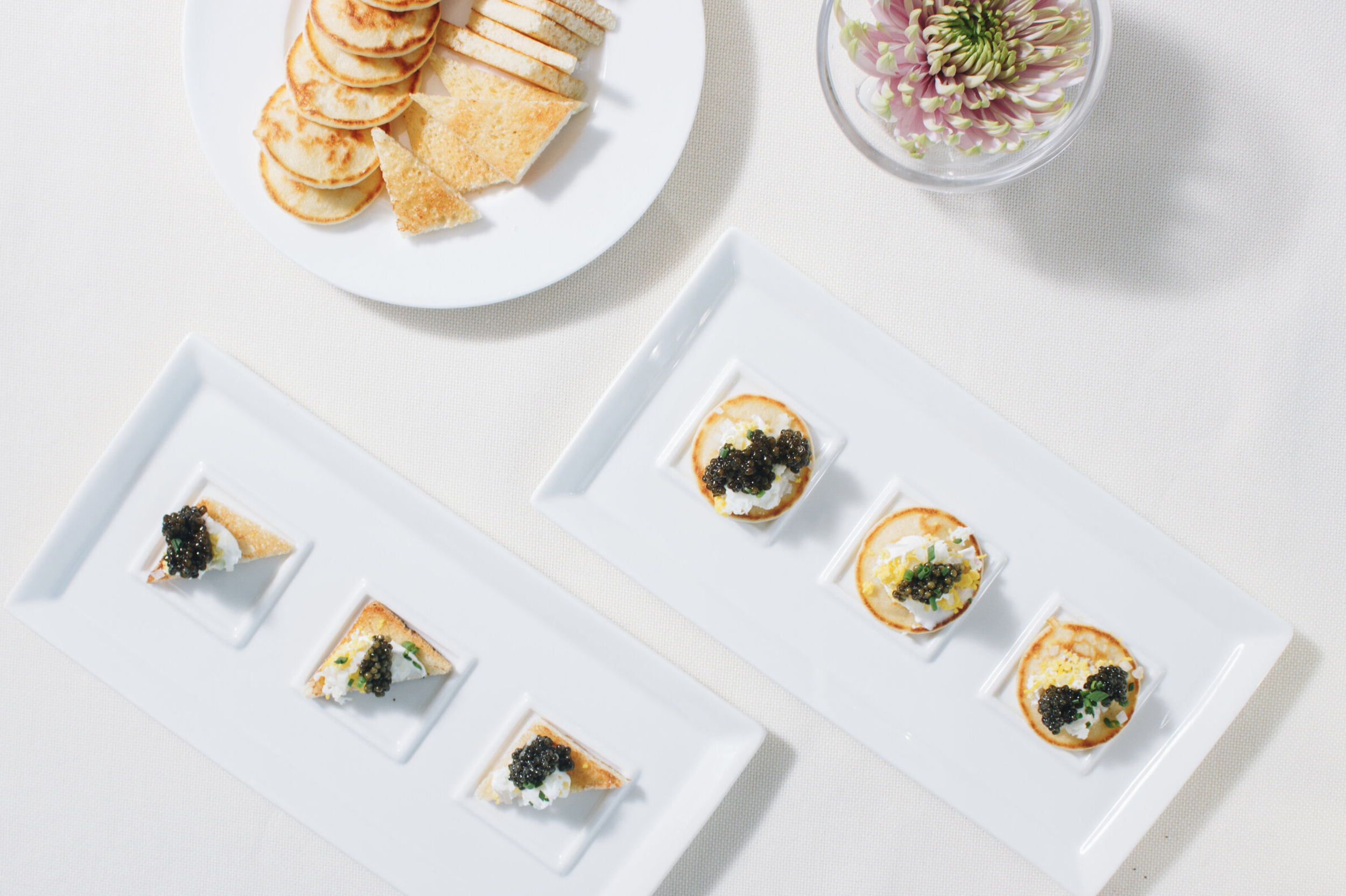 Appetizers on white ceramic plates with caviar, cheese, and crackers, and a bowl of flowers on a white tablecloth.