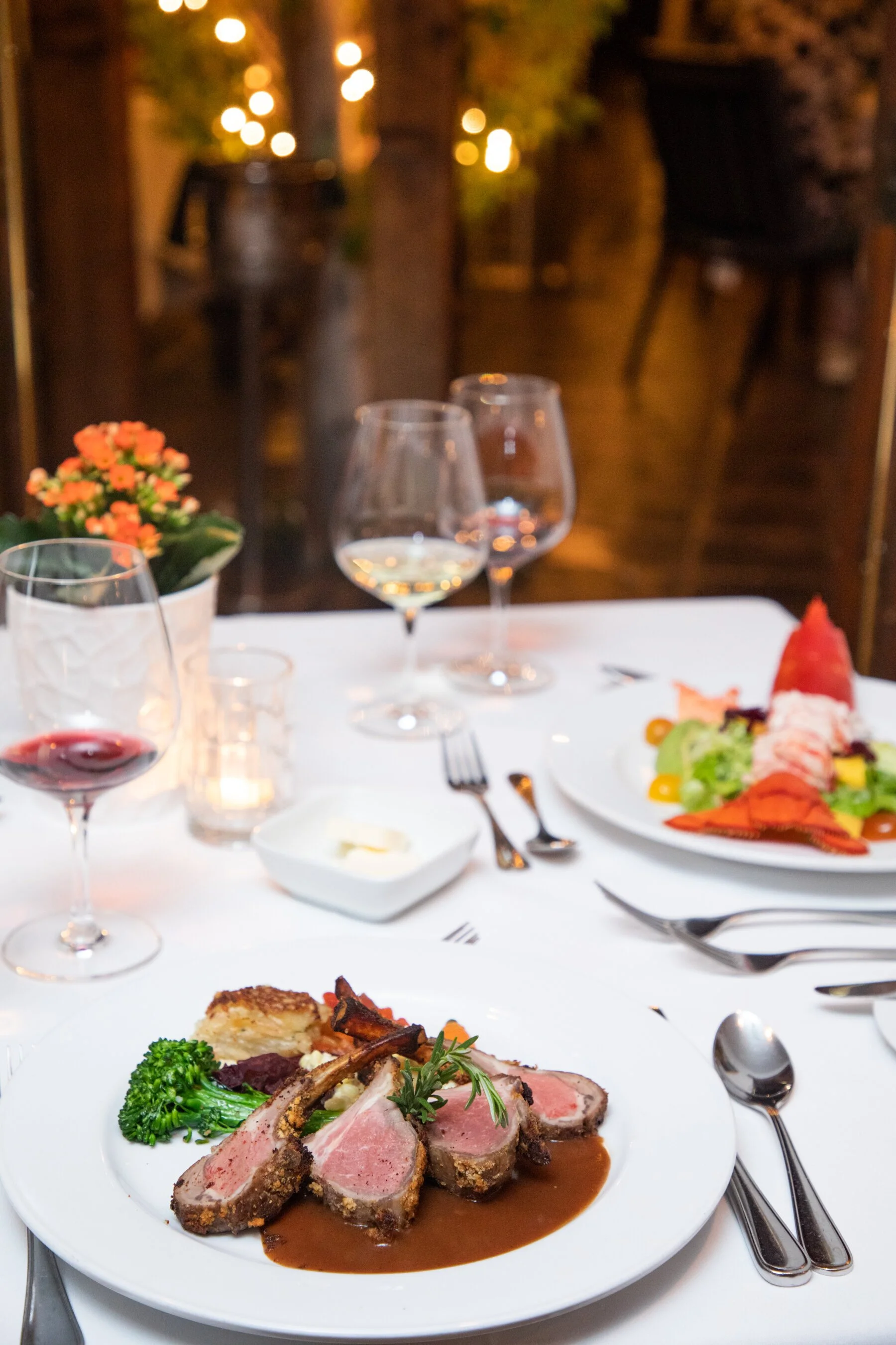 A formal dinner table set with red wine, white wine, a candle, and a flower arrangement, with a plate of lamb, vegetables, and mashed potatoes in the foreground, and a dessert plate with fruit and pastry in the background, all in a cozy, decorated di