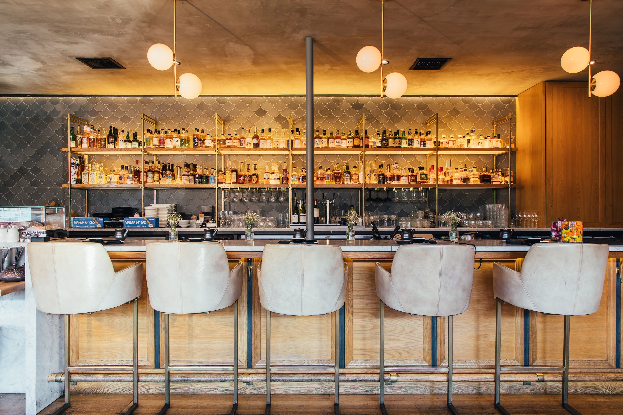Bar with beige chairs, illuminated shelves with various liquor bottles, wine glasses, and decorative flowers in a modern restaurant setting.