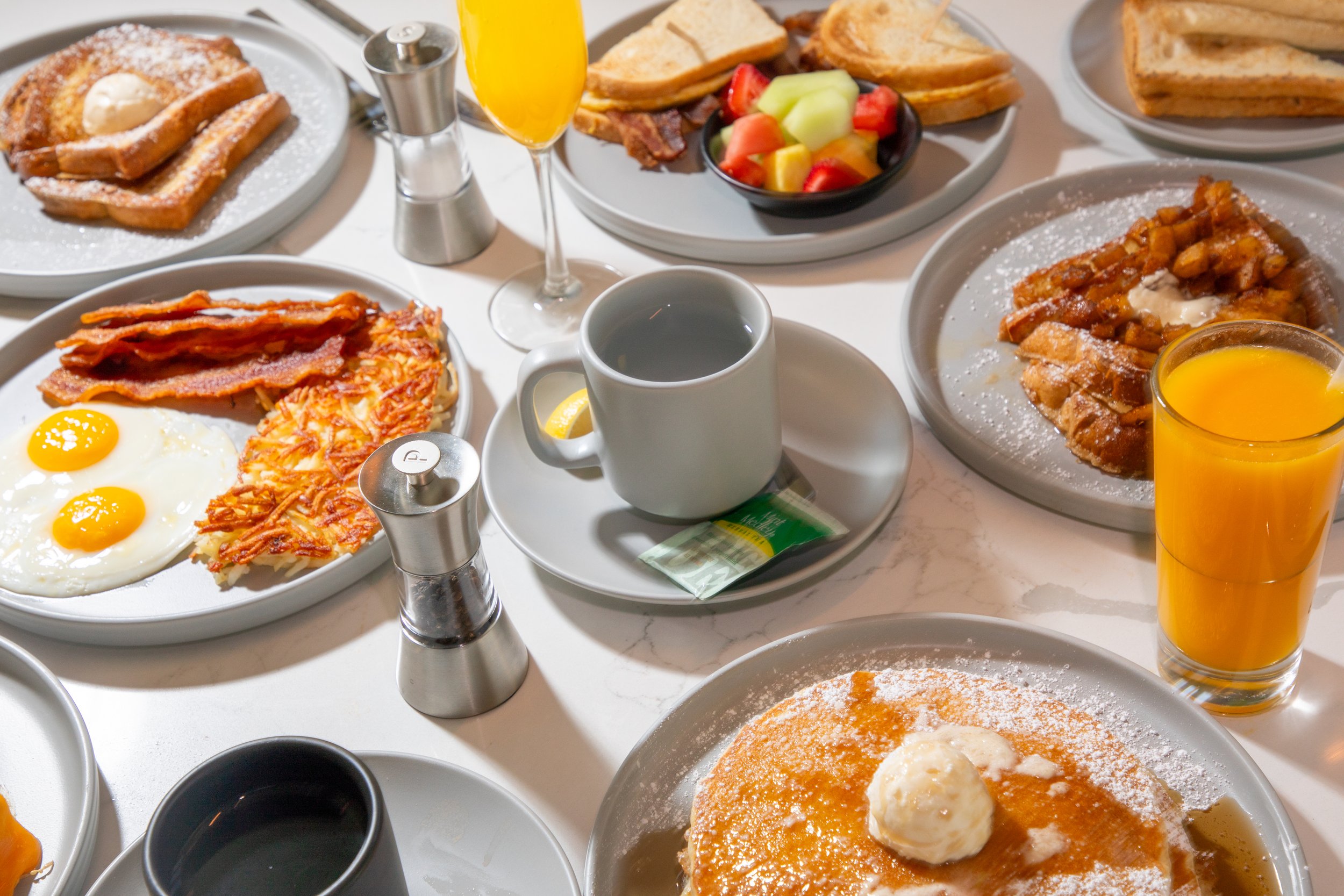 A breakfast spread including pancakes with butter, fried eggs, bacon, waffles with syrup, French toast, a fruit salad, orange juice, and coffee.