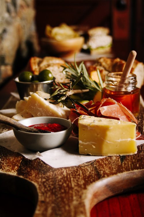 A cheese platter with various cheeses, including a yellow cheese, is served on a rustic wooden board with a small bowl of red jam, charcuterie, green olives, honey, and a sprig of rosemary. In the background, there are additional snacks or bread and 