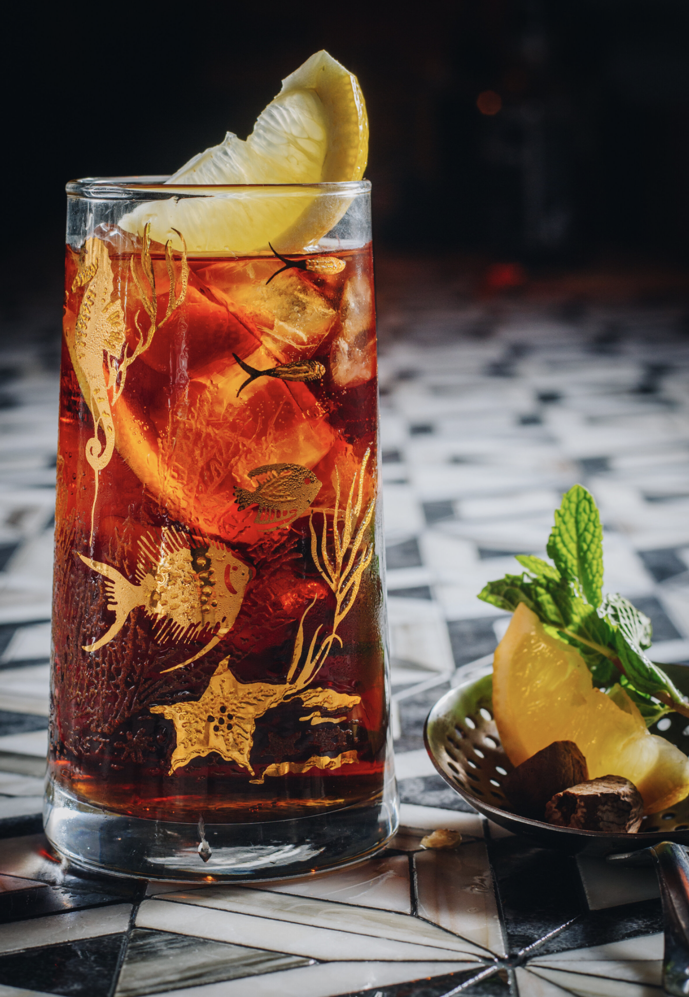 A tall glass of iced dark red beverage with lemon slice garnish and decorative sea life designs, placed on a table with a small dish of lemon, mint, and chocolate pieces.