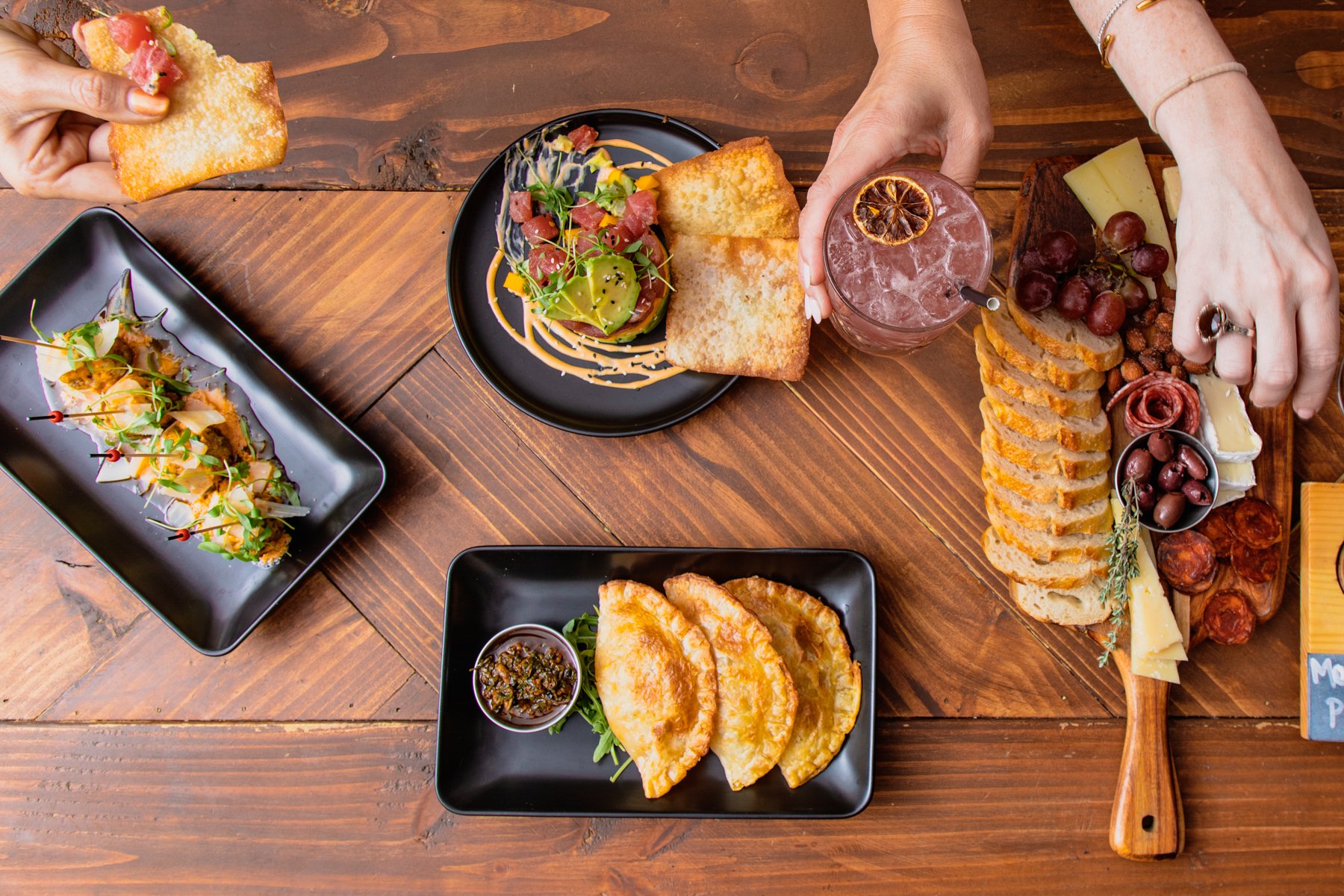 A top-down view of a table with various foods including a cheese and meat platter, fried dumplings with dipping sauce, a charcuterie board with sliced bread, cheese, grapes, and meats, and small appetizers. A person reaches for the charcuterie board 