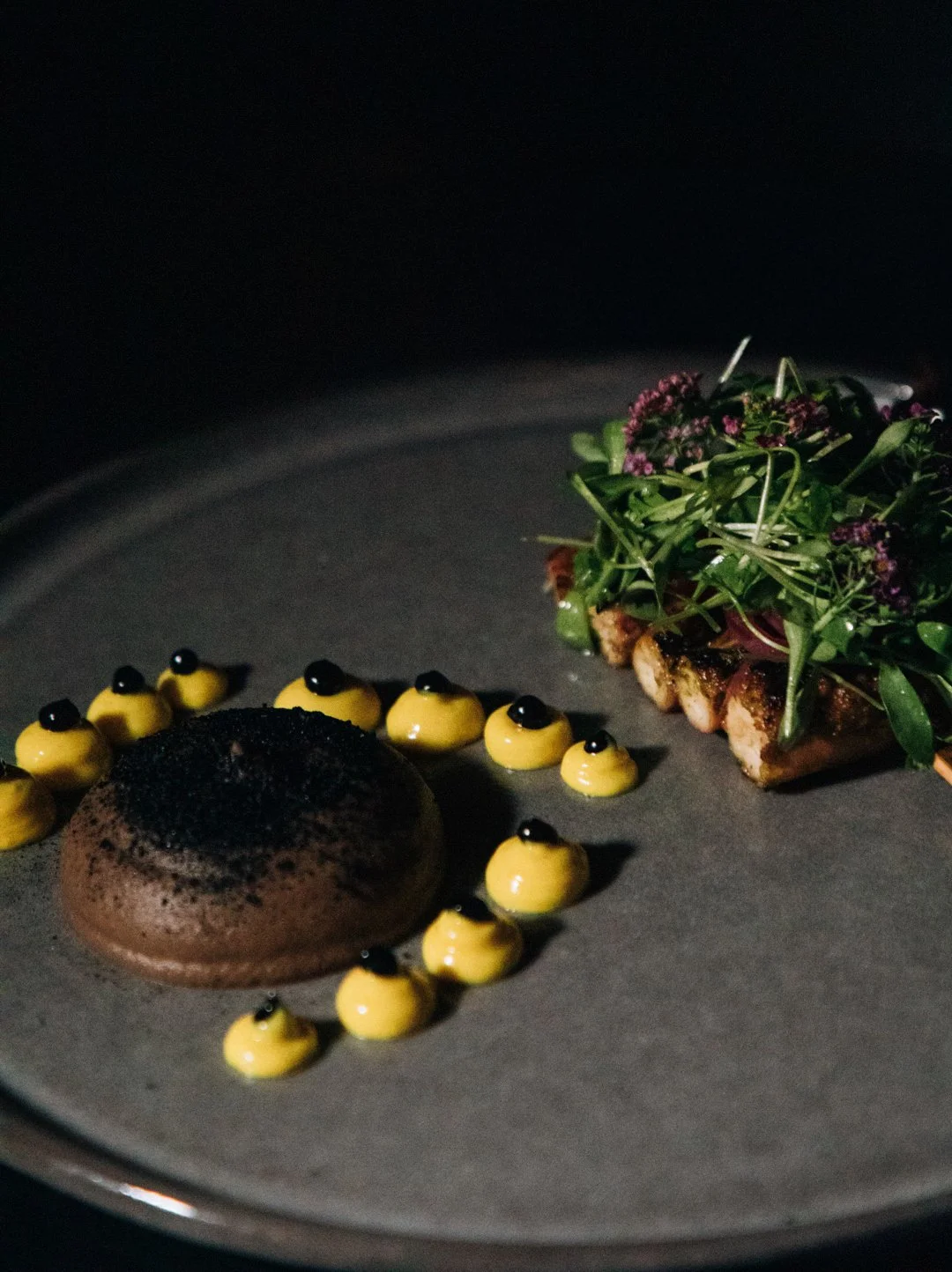 A gourmet dish featuring a round chocolate dessert garnished with black pearls and yellow sauce, served alongside a piece of toast topped with greens and edible flowers.