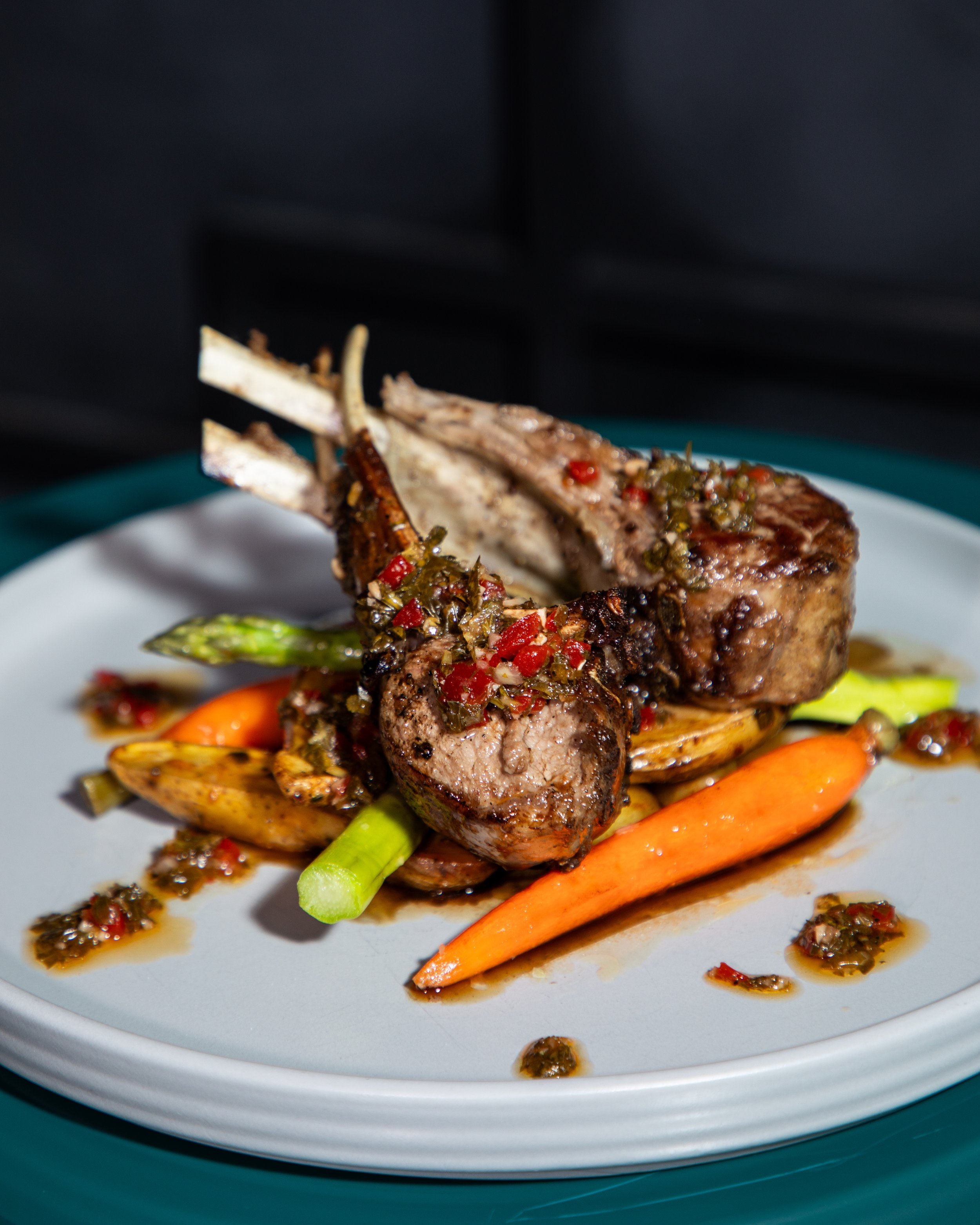 Grilled lamb chops with herbs and chili sauce, served with roasted carrots, asparagus, and potatoes on a white plate.
