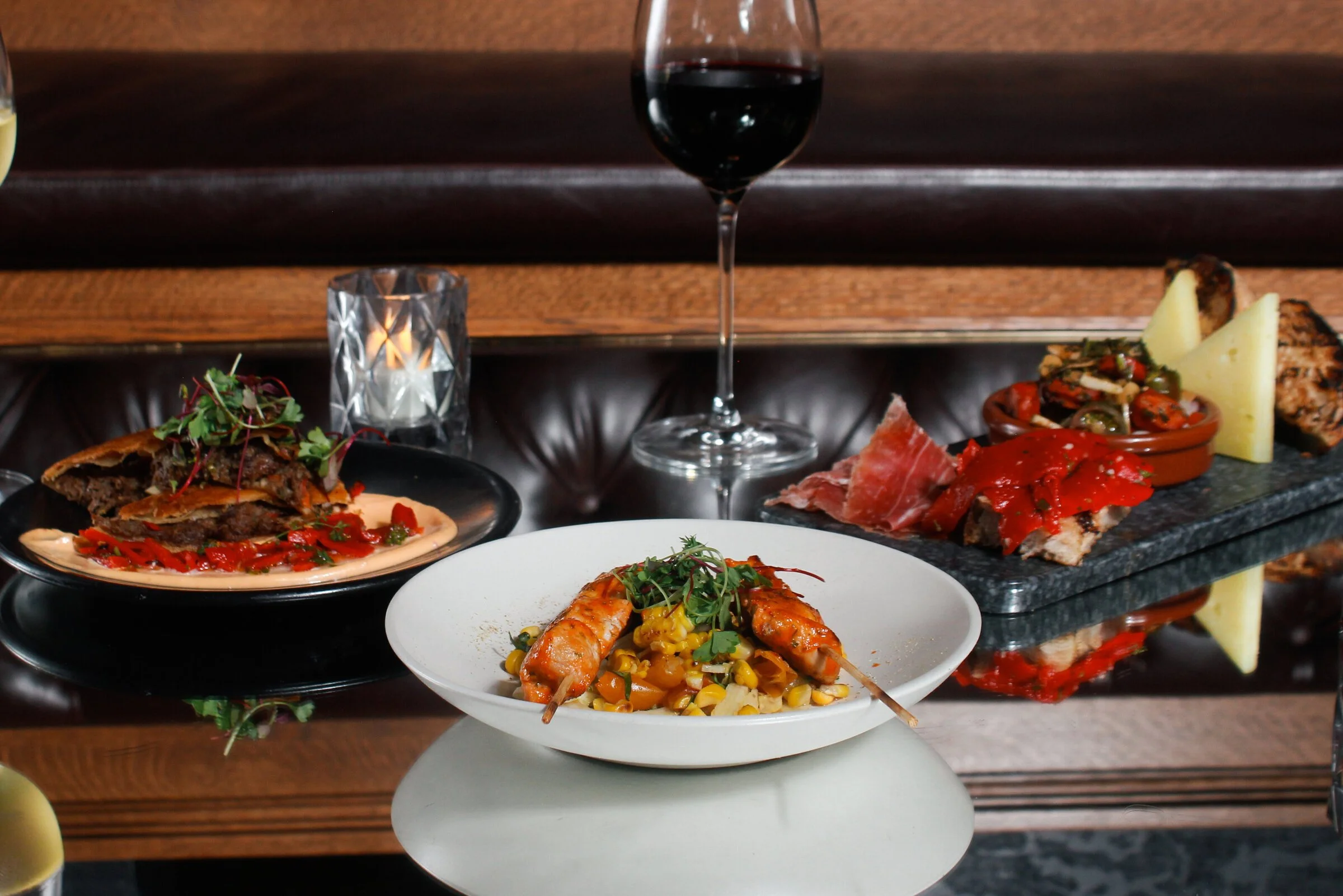A glass of red wine, a candle holder, and an assortment of appetizers including grilled meats, cheeses, bread, and seafood on a reflective glass table.