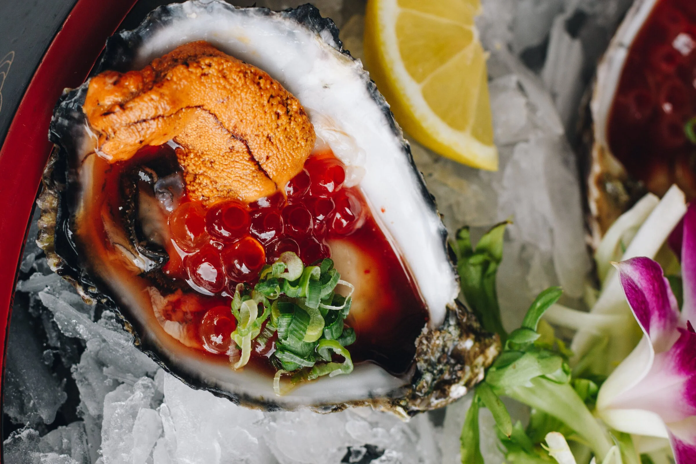 A fresh oyster on the half shell served on a bed of ice, garnished with green onions, red roe, and a lemon wedge in the background.