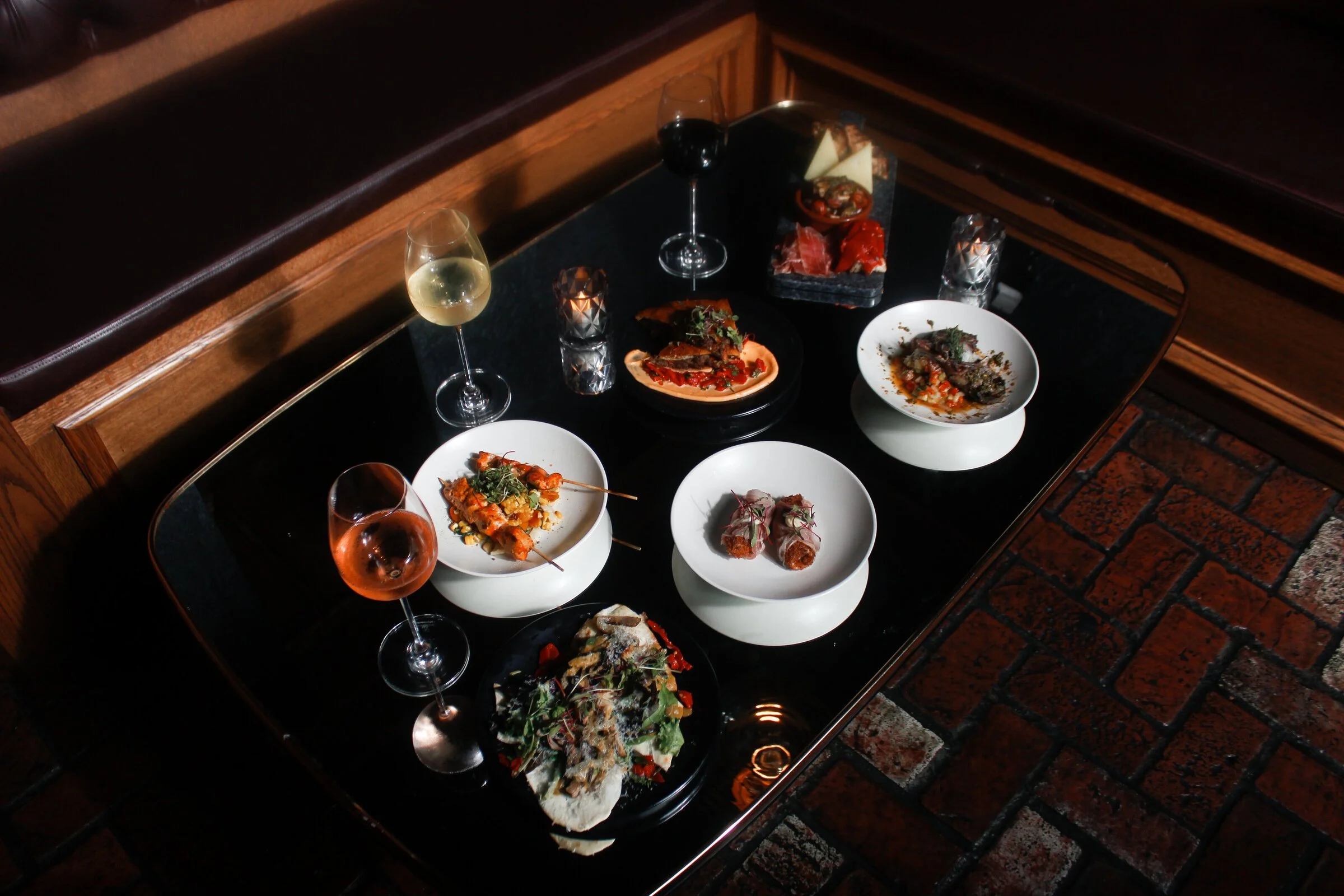A top-down view of a black table with various dishes and drinks, including plates of food, wine glasses with white, red, and rosé wine, and a small dish of charcuterie, set on a brick floor.
