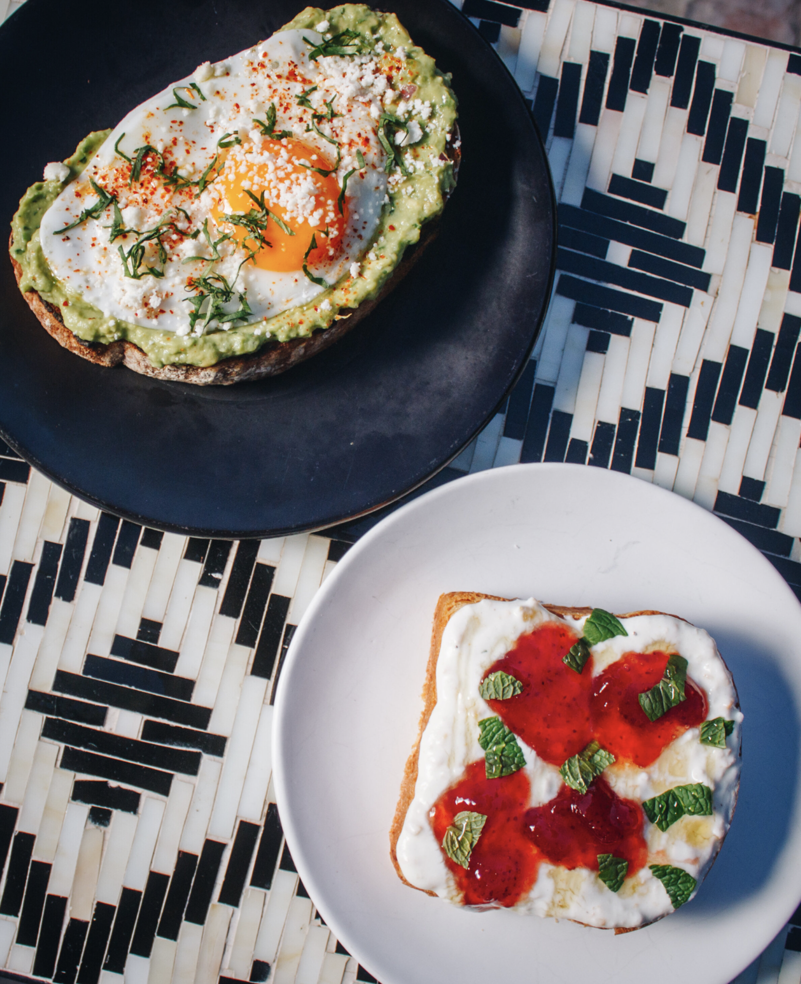 Open-faced avocado toast with a fried egg, chopped herbs, and cheese on dark plate; American-style toast with white cheese, red fruit preserves, and fresh mint on white plate.