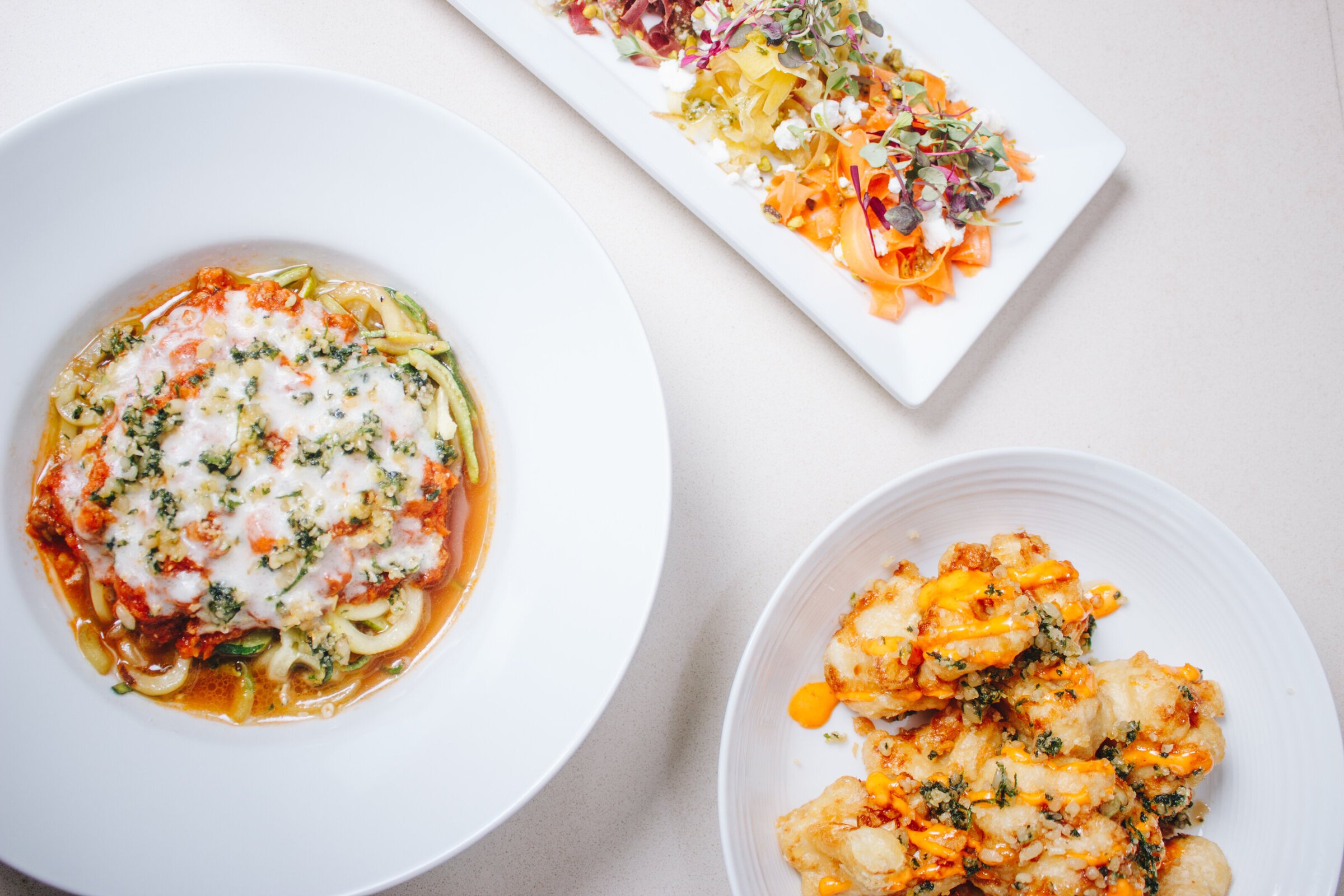 Three plates of food on a light-colored surface: a bowl of cheesy pasta with vegetables, a rectangular plate of colorful salad with microgreens, and a plate of fried cauliflower with sauce.