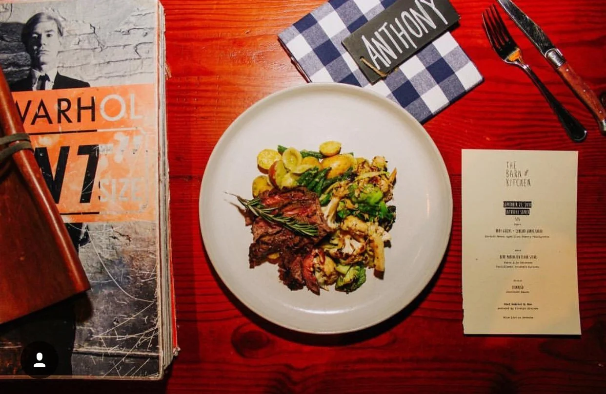 A white plate with cooked vegetables and a roast beef with a sprig of rosemary in the center on a red wooden table. Surrounding items include a book, a  napkin with the name 'Anthony,' a fork and knife, and a menu from 'The Barn & Kitchen.'