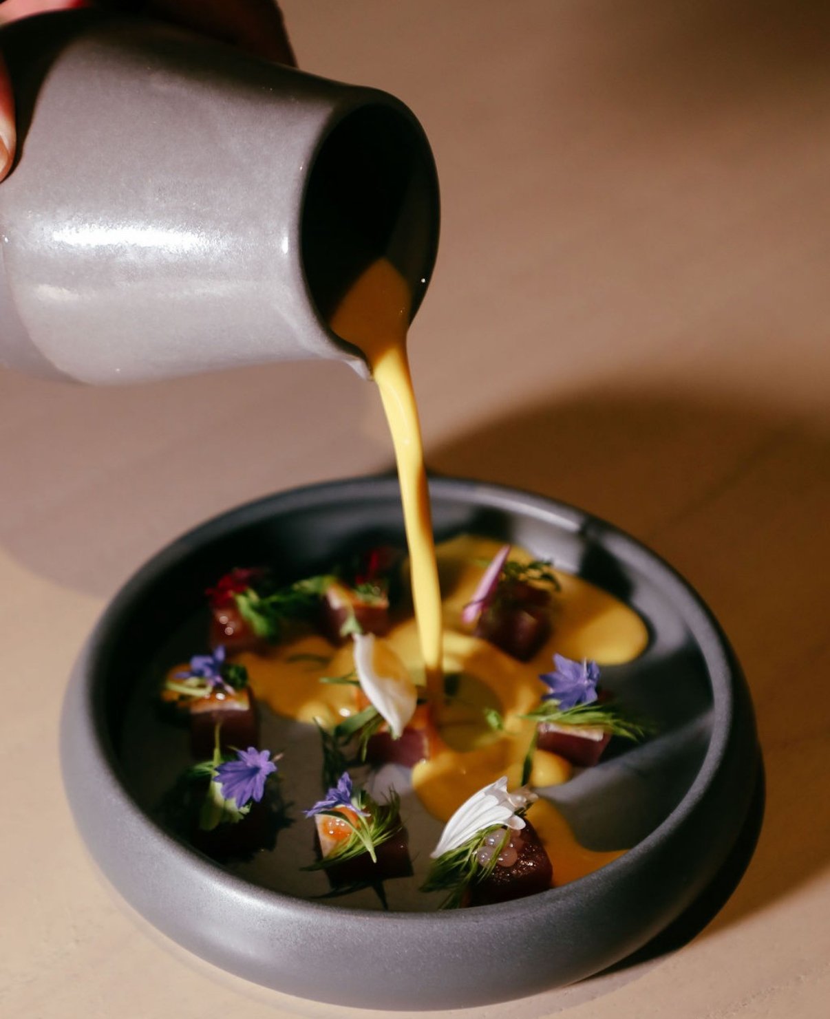 Pouring yellow soup or sauce onto a black bowl with flower petals and small pieces of meat or vegetables.