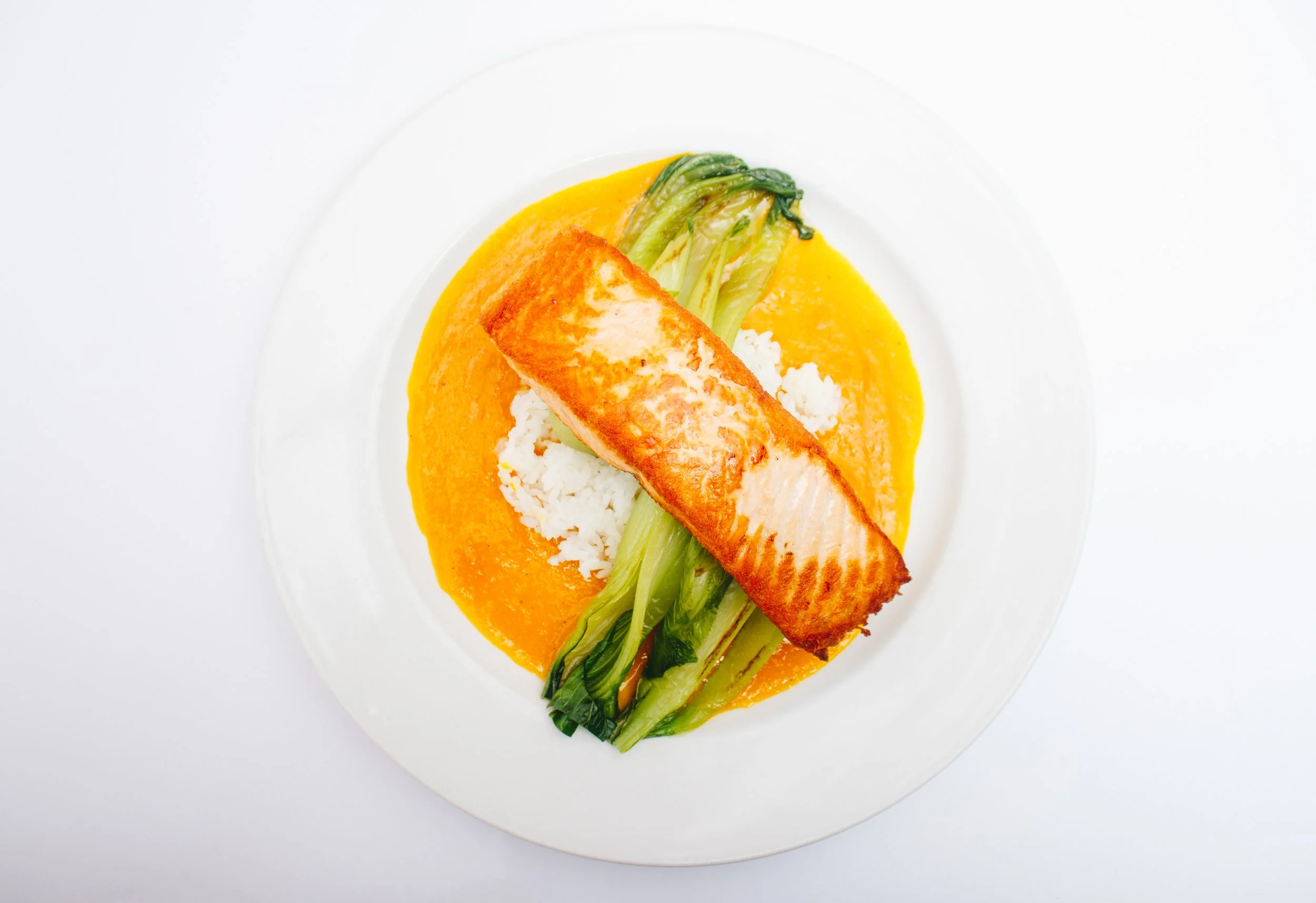 A white plate with a cooked salmon fillet, white rice, sautéed bok choy, and a yellow sauce on a white background.