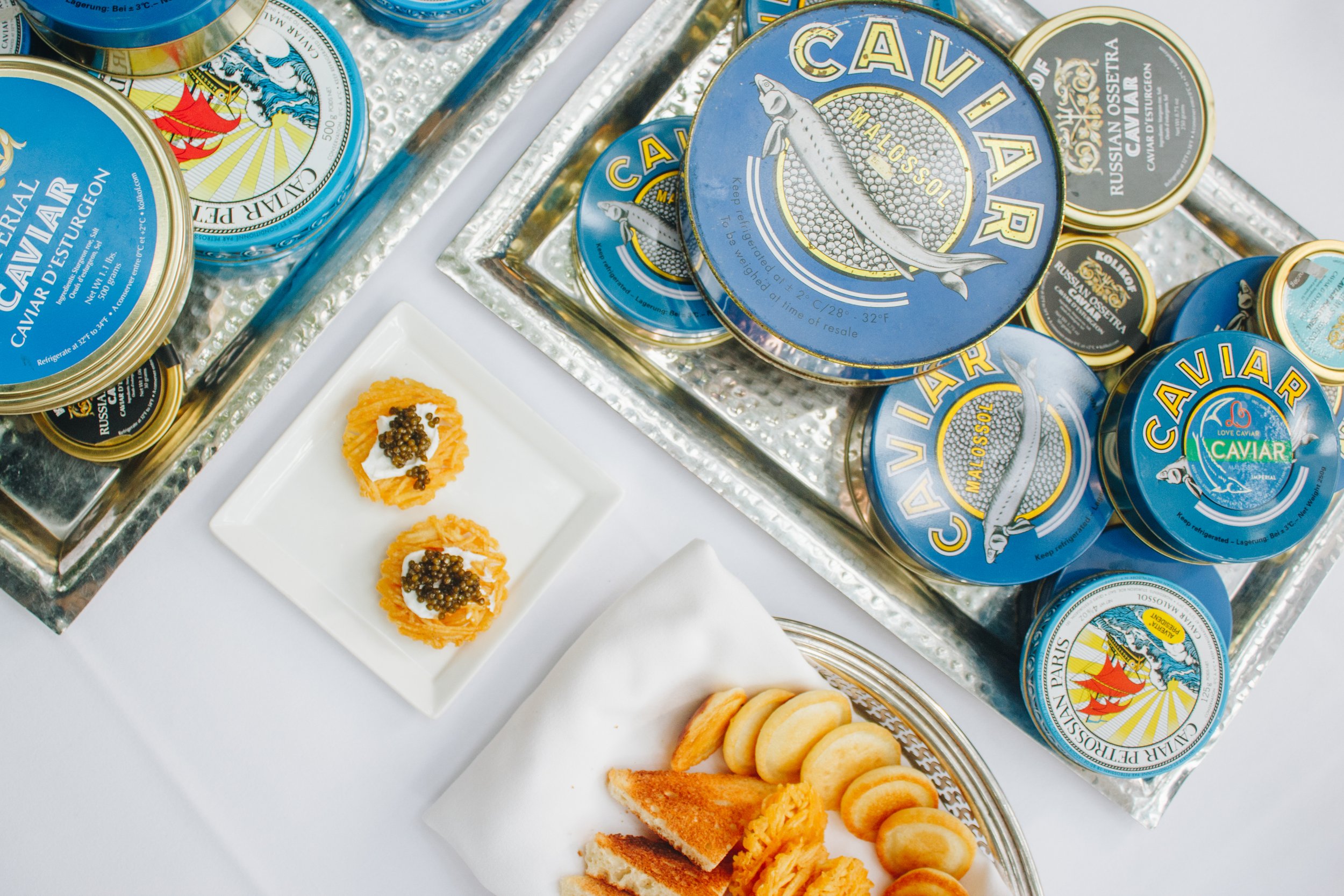 A display of various tins of caviar, with some served on a white plate alongside bread, crackers, and garnishes, on a silver tray and white table.