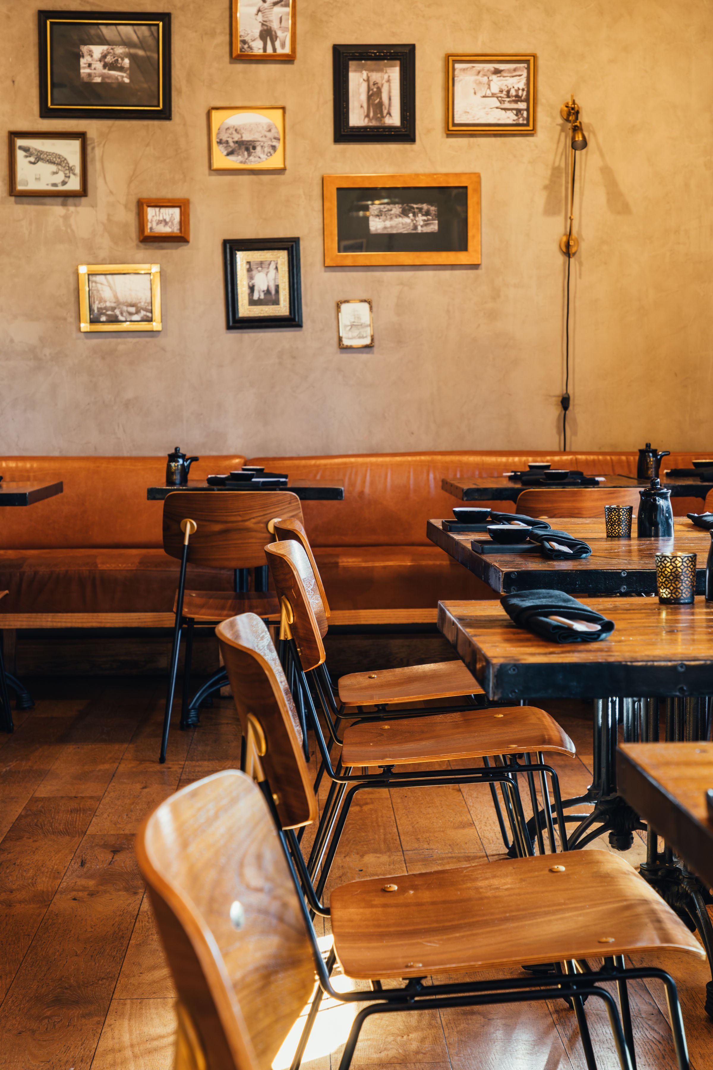 Empty restaurant with wooden chairs, tables set with black napkins and condiments, a beige wall decorated with framed black and white photos, and a wall-mounted lamp.