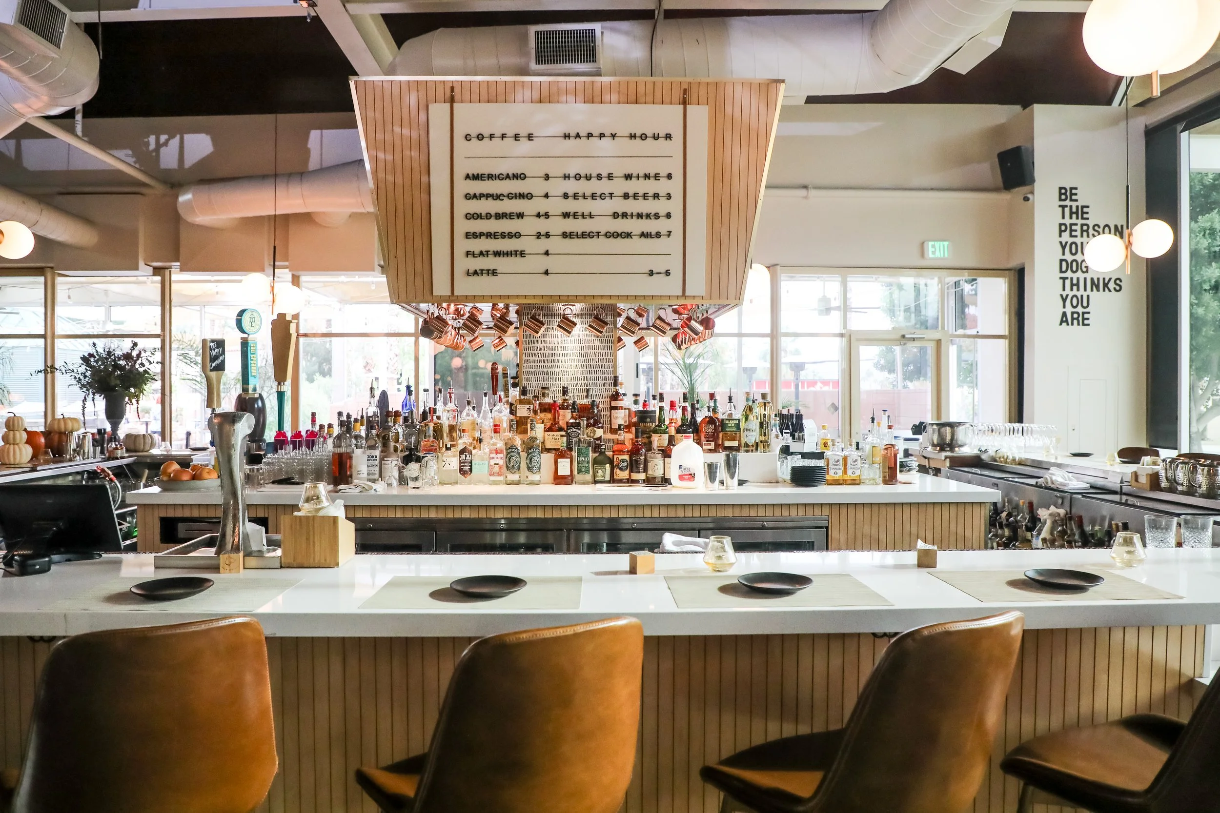 Interior of a modern bar and restaurant with a white countertop and brown leather chairs, a variety of liquor bottles behind the bar, a large menu board hanging above, decorative hanging mugs, and large windows providing natural light.