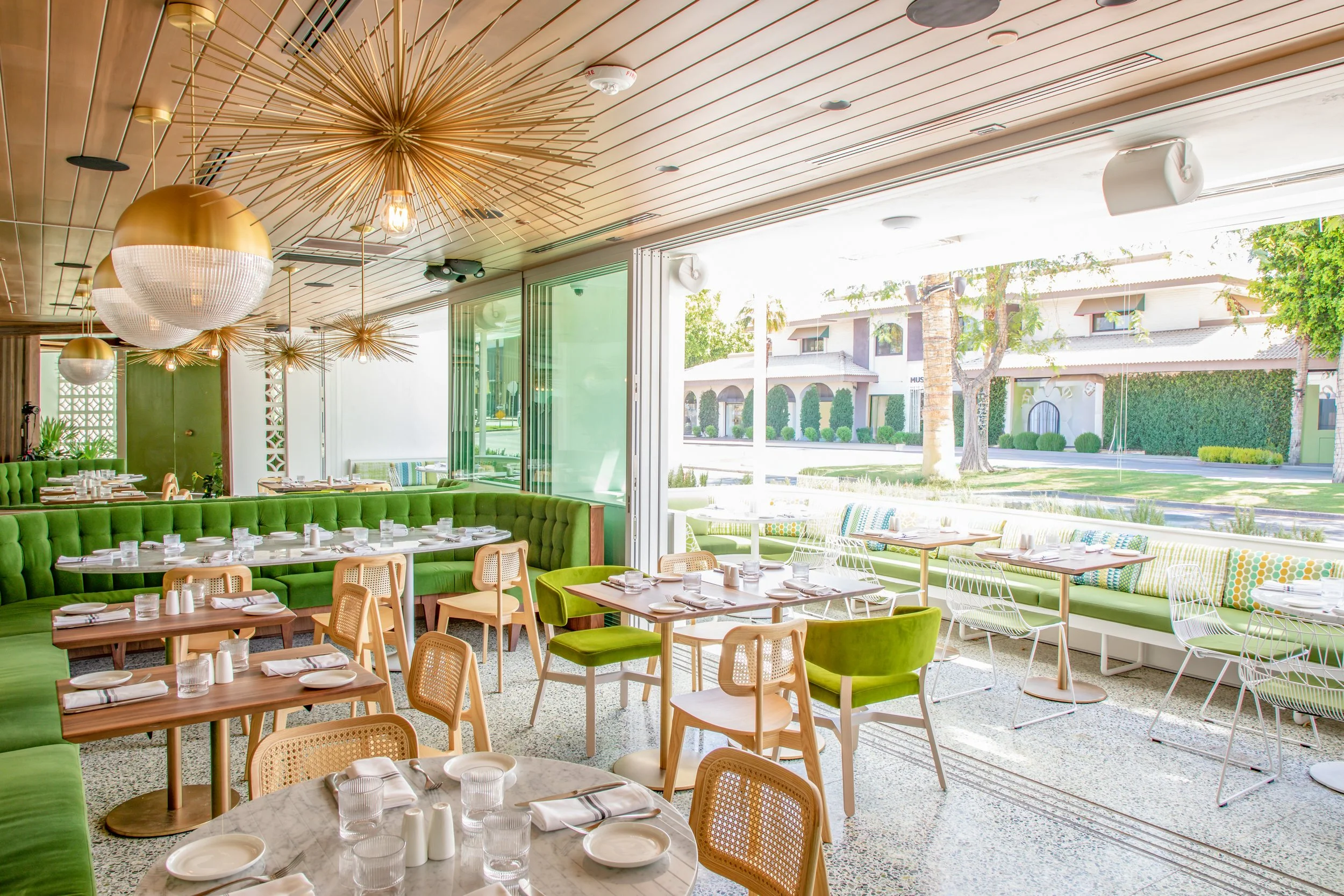 Bright, modern restaurant interior with green seating, wooden tables, and large windows showing a sunny outdoor street view with trees and residential buildings.
