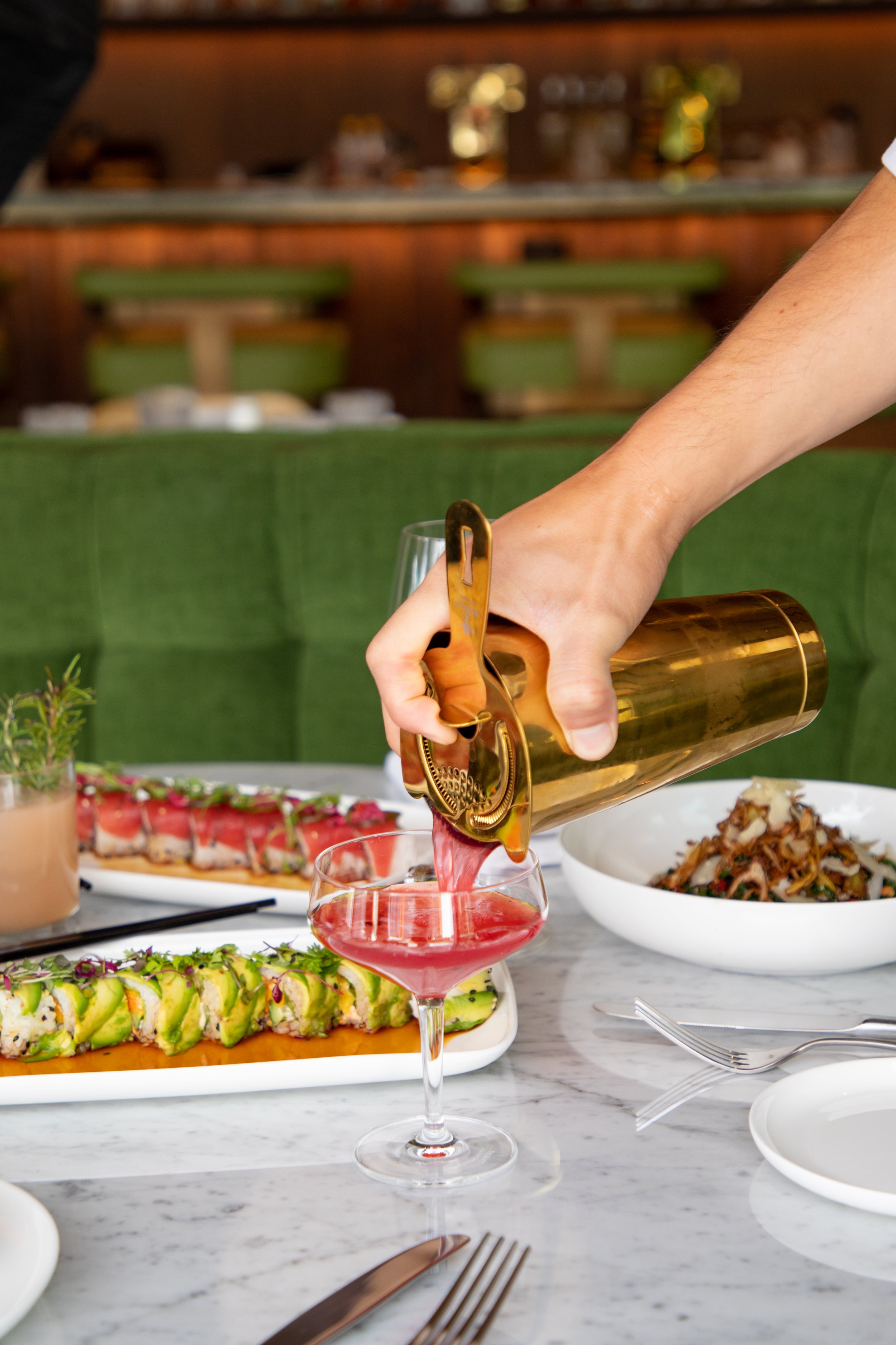 Person pouring pink-colored drink into a wine glass at a table with sushi and other dishes in a restaurant.