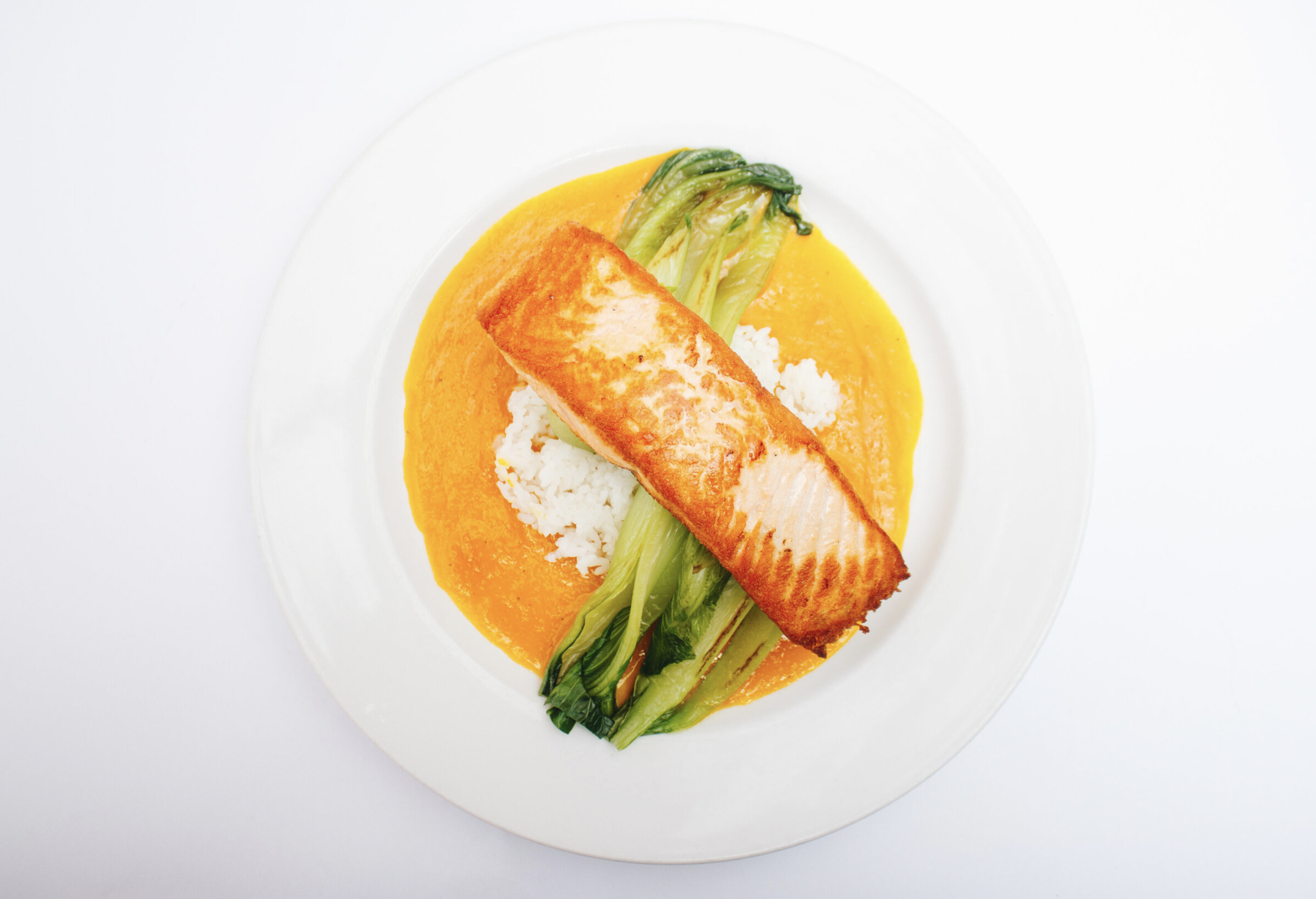 Seared fish fillet served with a bed of white rice, green bok choy, and a yellow sauce on a white plate.