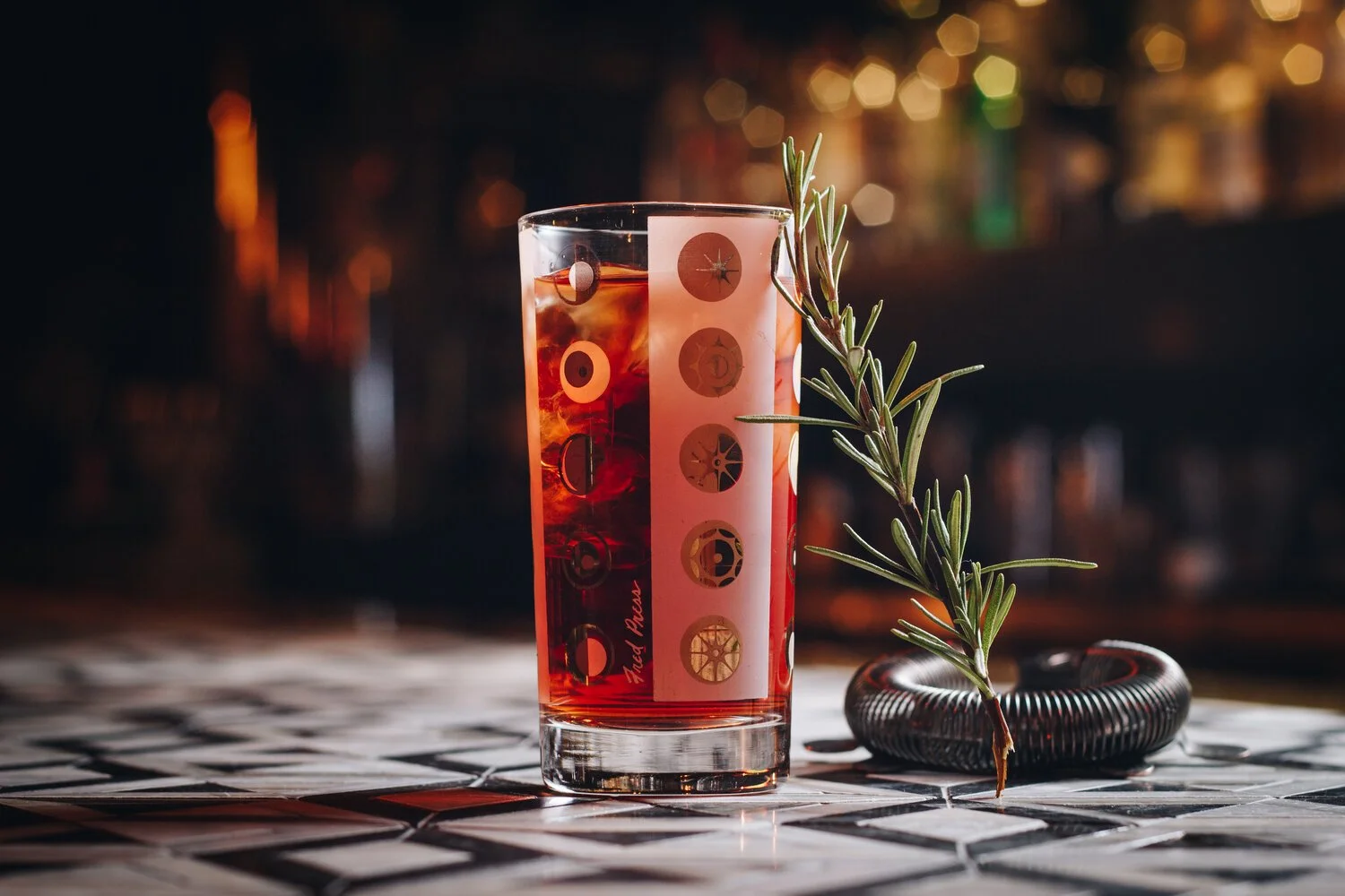 A tall glass of red cocktail with ice, garnished with a sprig of rosemary, sitting on a tiled surface with a blurred bar background.