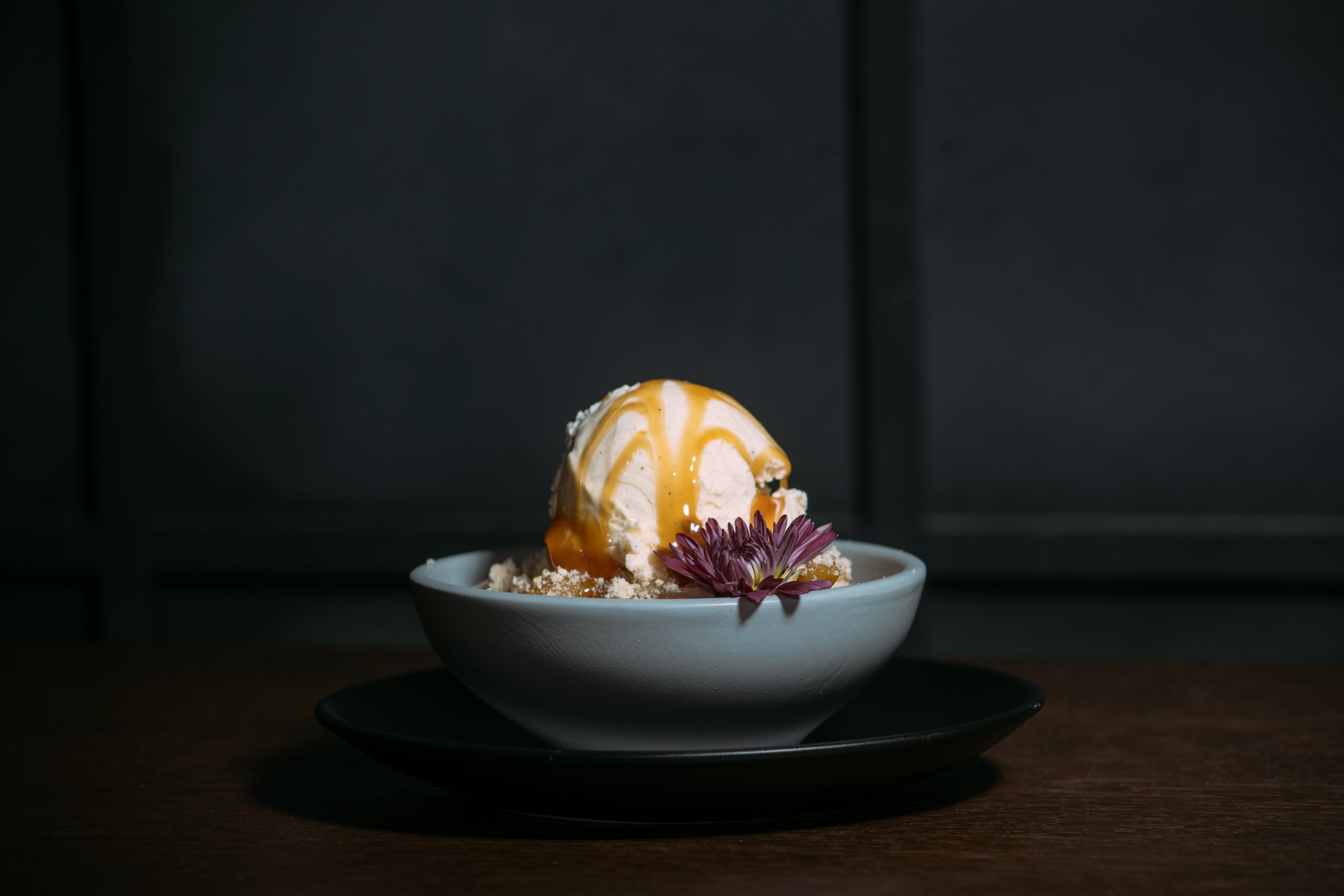 A bowl of ice cream with caramel sauce, garnished with a purple flower on a black plate on a wooden table against a dark background.