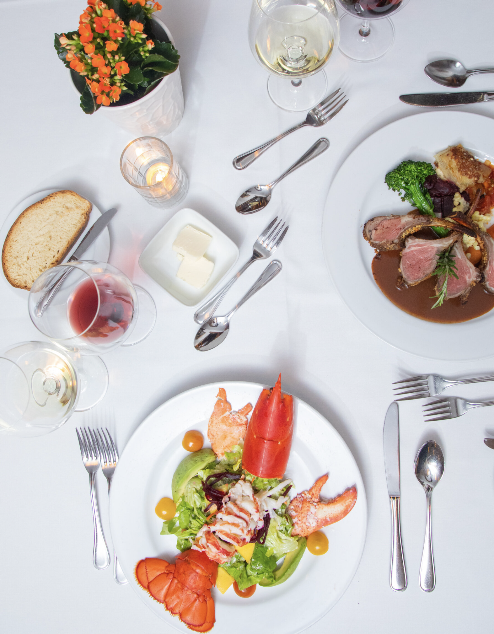 A top-down view of a formal dining table set with wine glasses, a lobster salad, a plate of sliced beef with vegetables, bread, butter, and a small potted plant, with silverware and lit candles.