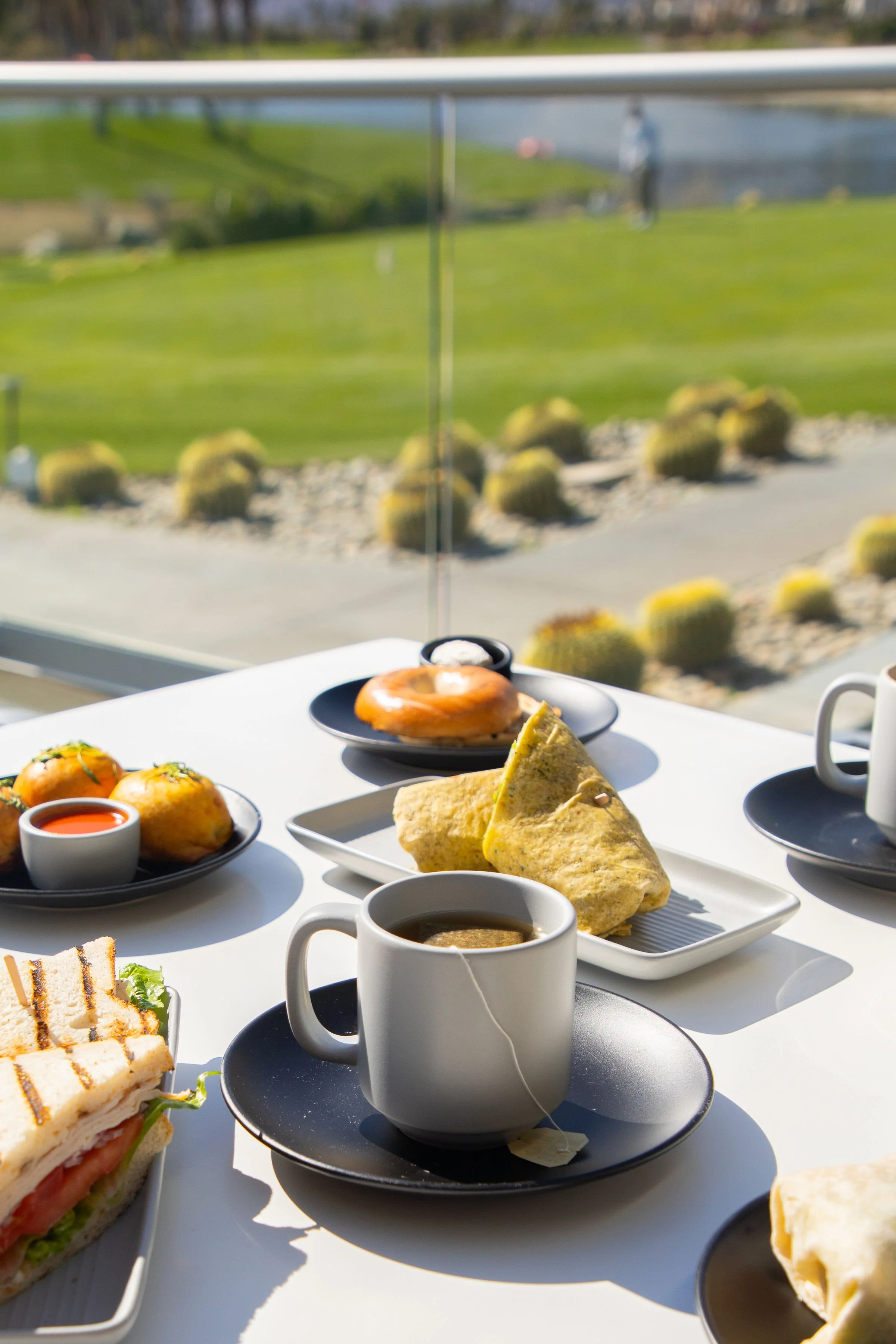 Breakfast spread with a coffee cup, sandwich, rolls, and dips on a table near a window overlooking a landscaped outdoor area.