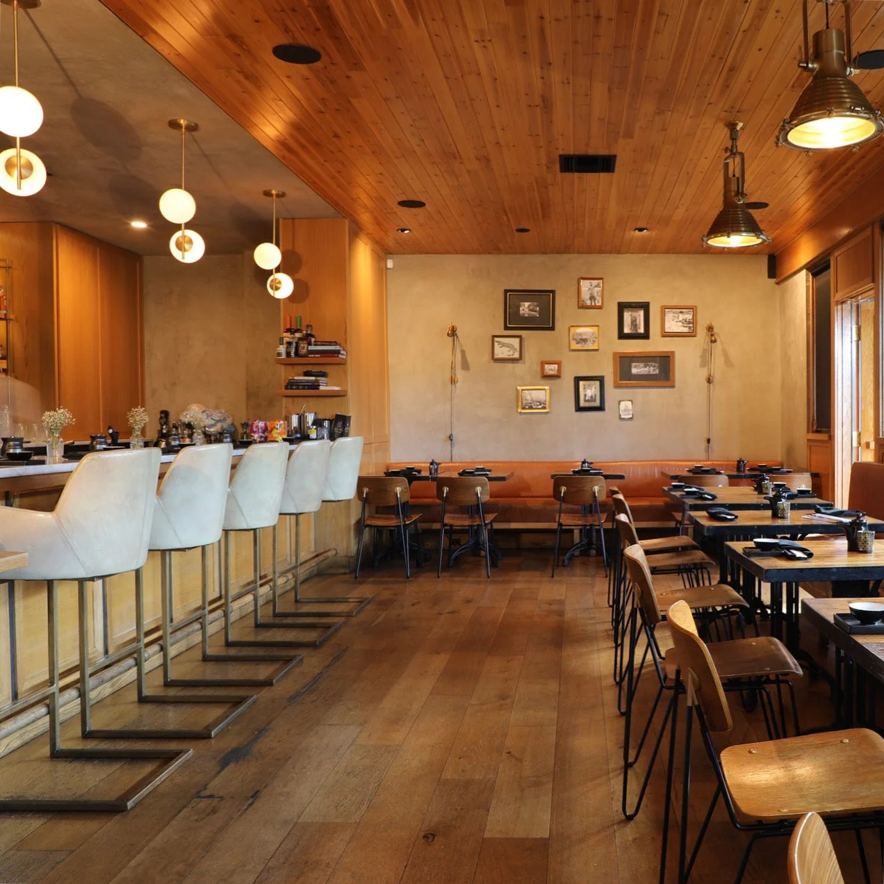 Interior of a cozy restaurant with wood-paneled ceiling and floor, featuring a bar area on the left with white bar stools, and dining tables on the right with black chairs, plates, utensils, and glasses. The wall displays a gallery of framed pictures