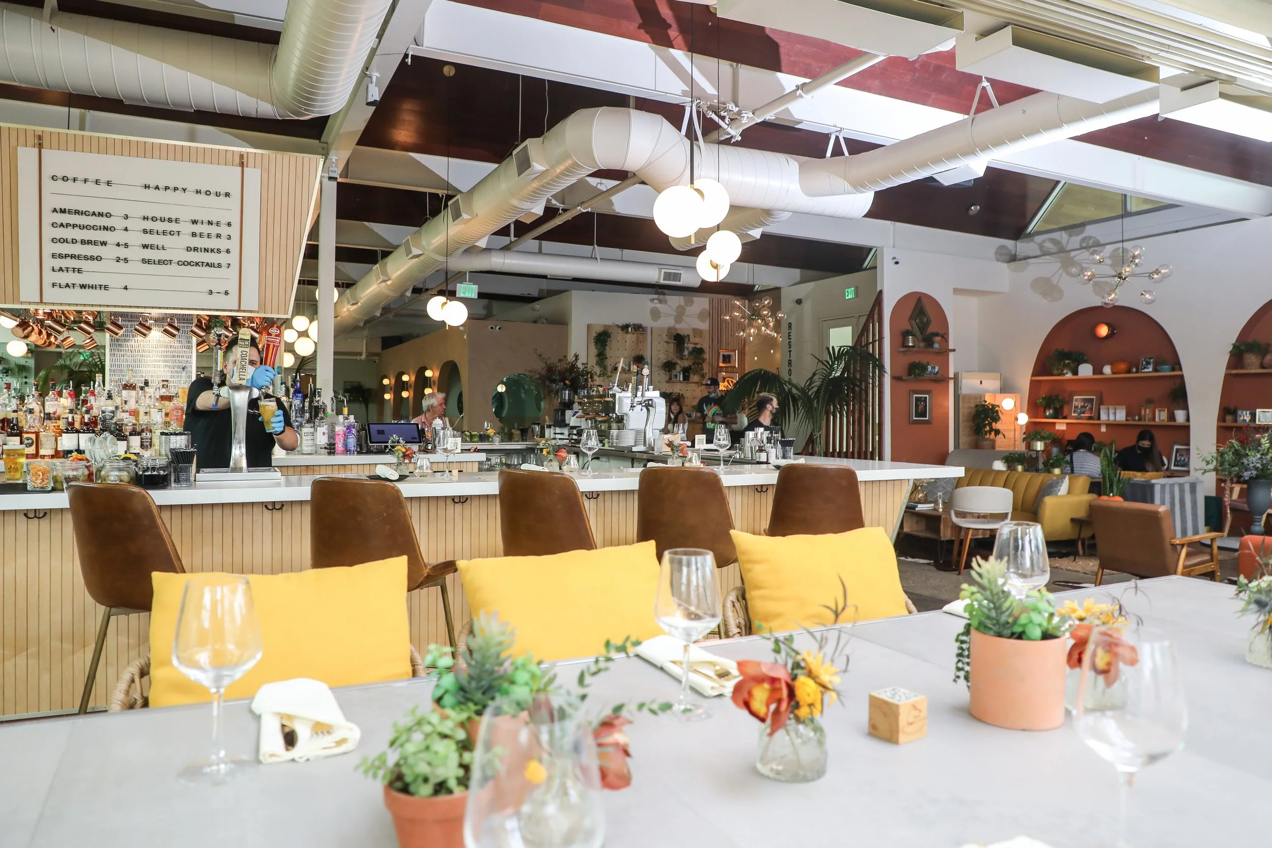 A stylish cafe interior with a bar, seating area, and decorative plants. The bar has a white countertop with bar stools, and a bartender pouring drinks. The seating includes yellow pillows and various chairs, with a menu board visible above. The ambi