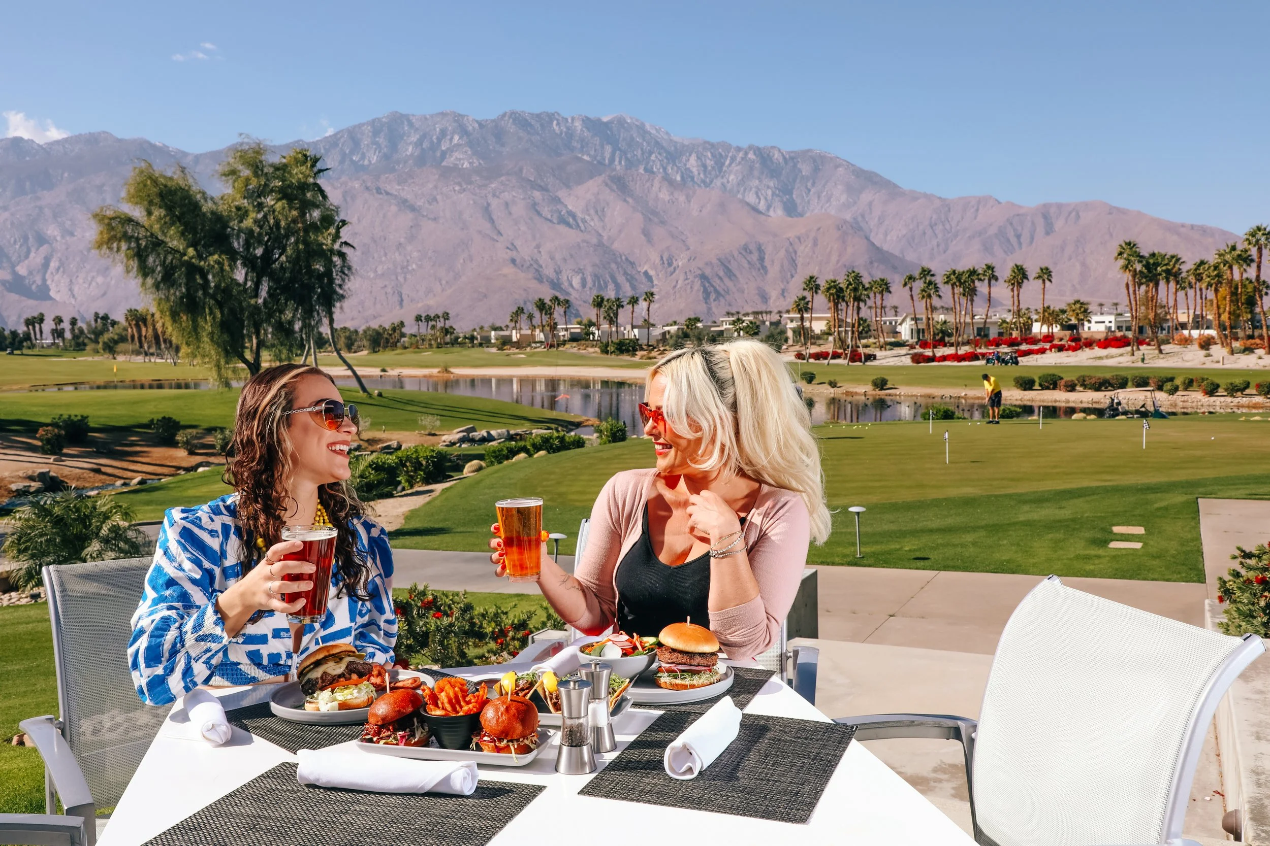 Two women enjoying drinks and food at an outdoor table near a golf course with mountains in the background