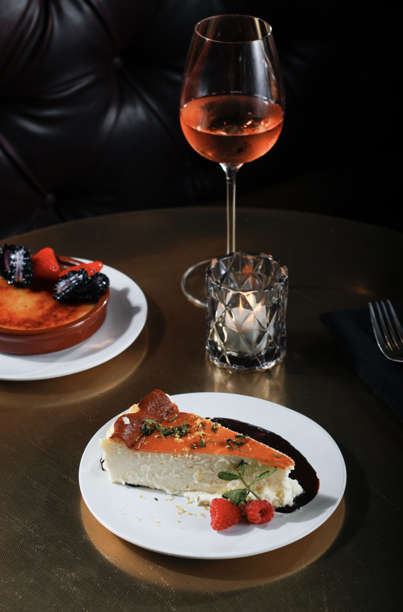 A dessert plate with a slice of cheesecake garnished with raspberries and a sprig of mint, a glass of rosé wine, and a patterned candle holder on a dark table.