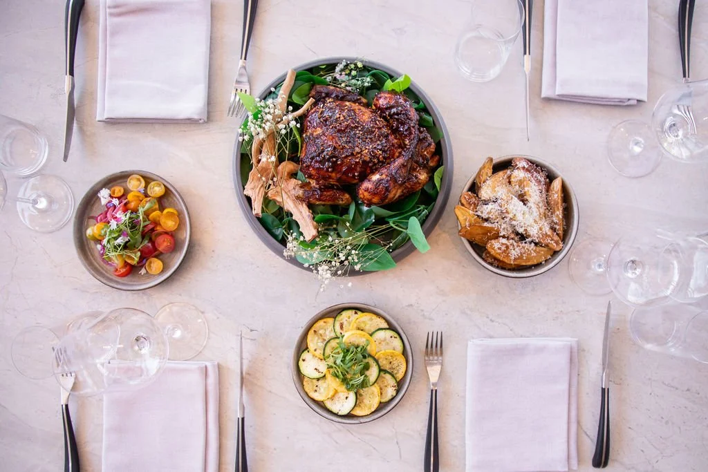 A table with a roasted chicken garnished with greens and flowers, surrounded by bowls of salad, potato wedges with grated cheese, and sliced zucchini, set with white napkins, cutlery, and empty wine glasses.