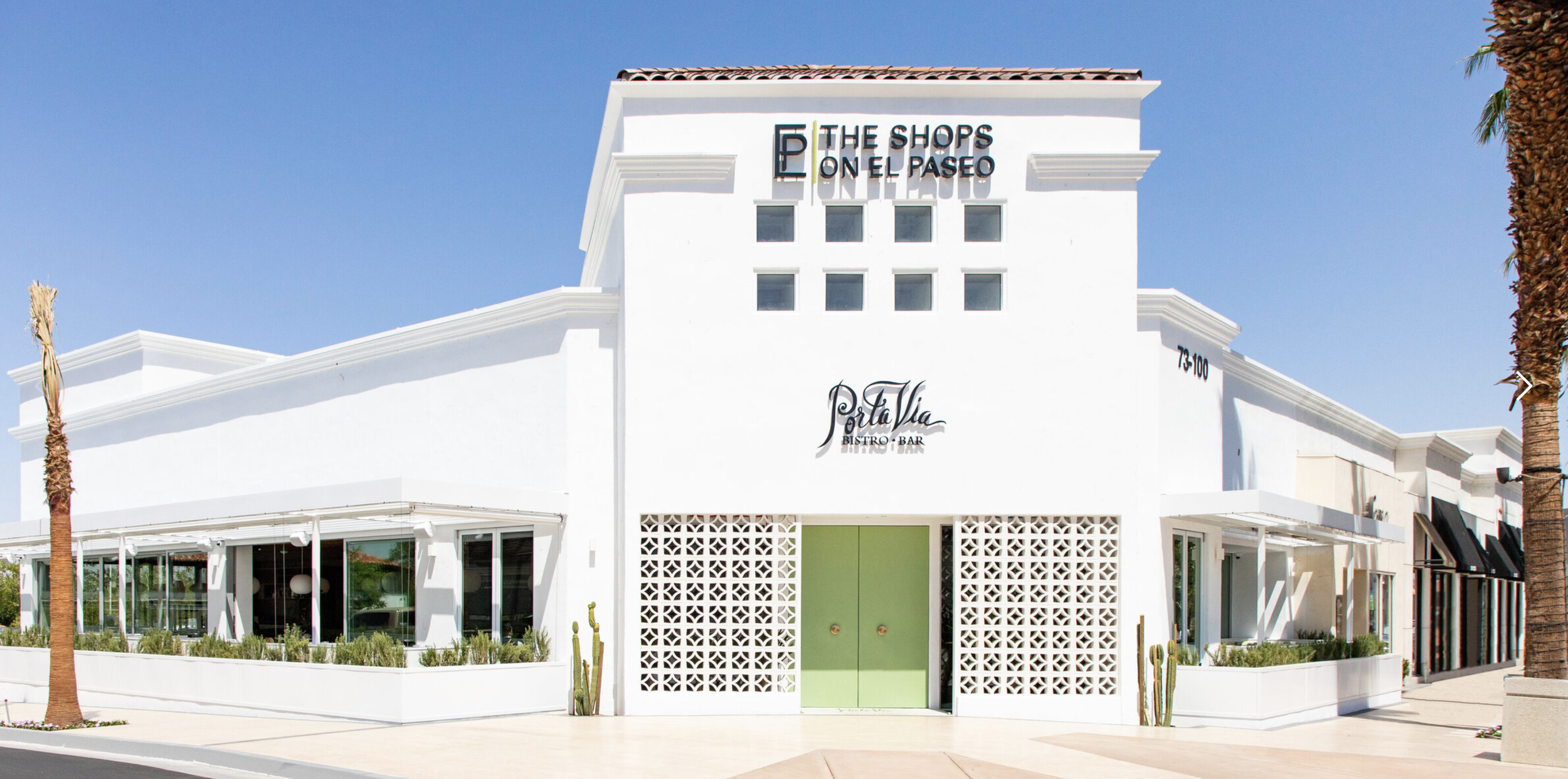 White shopping complex with sign 'The Shops on El Paseo' and a restaurant sign 'Porta Vino Bistro + Bar' on front, flanked by palm trees under a clear blue sky.