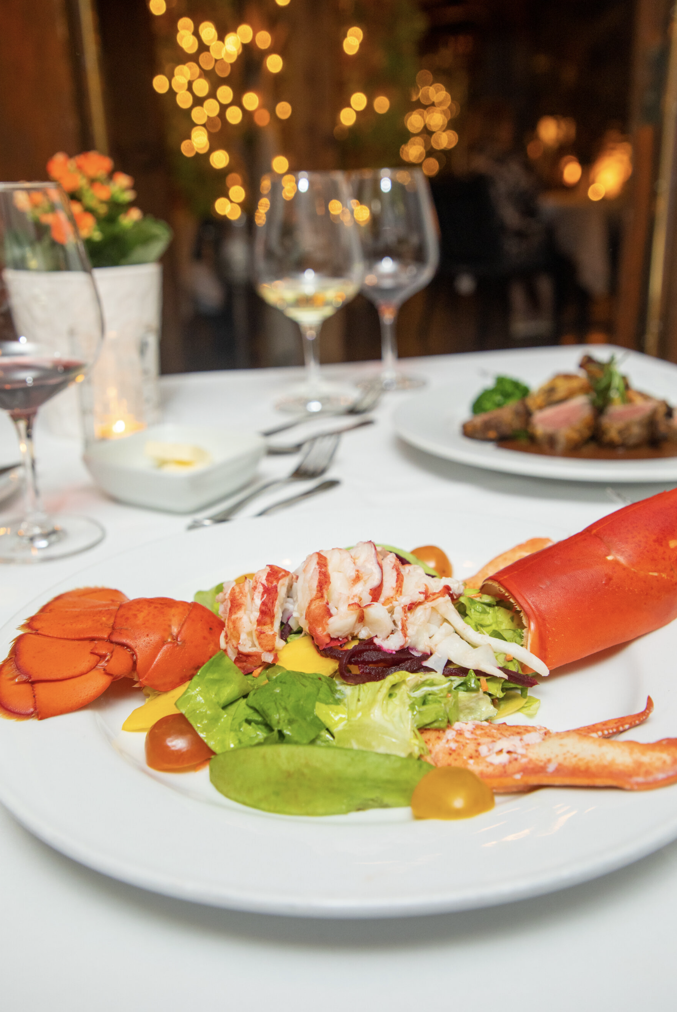 Lobster on a bed of salad with various vegetables, served on a white plate, with a table set for dining in the background.