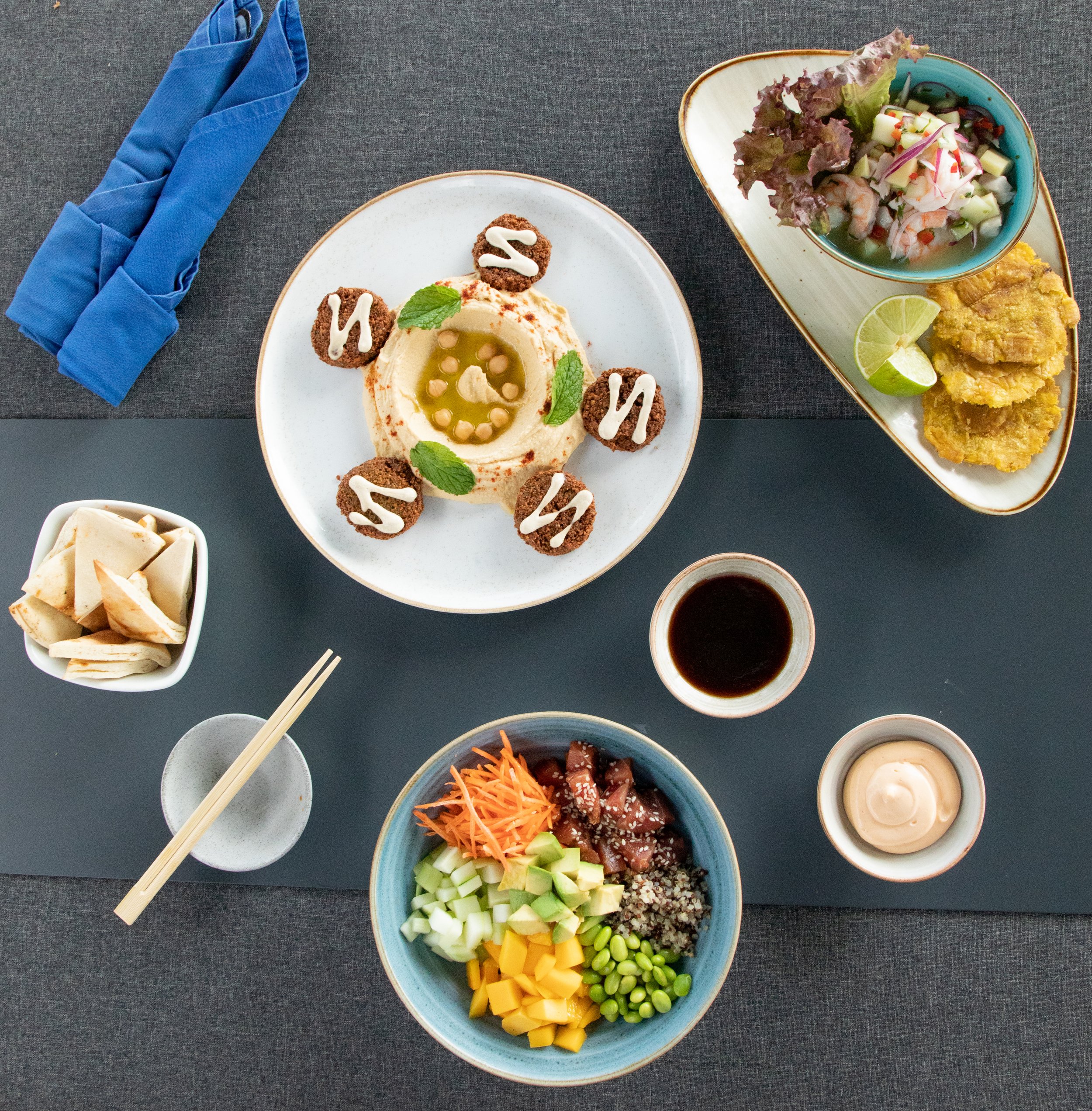 A variety of dishes on a table including a bowl of mixed vegetables and raw fish, a plate of hummus garnished with olive oil and herbs, small bowls of soy sauce, creamy dressing, and sliced bread, with chopsticks resting on a small dish.