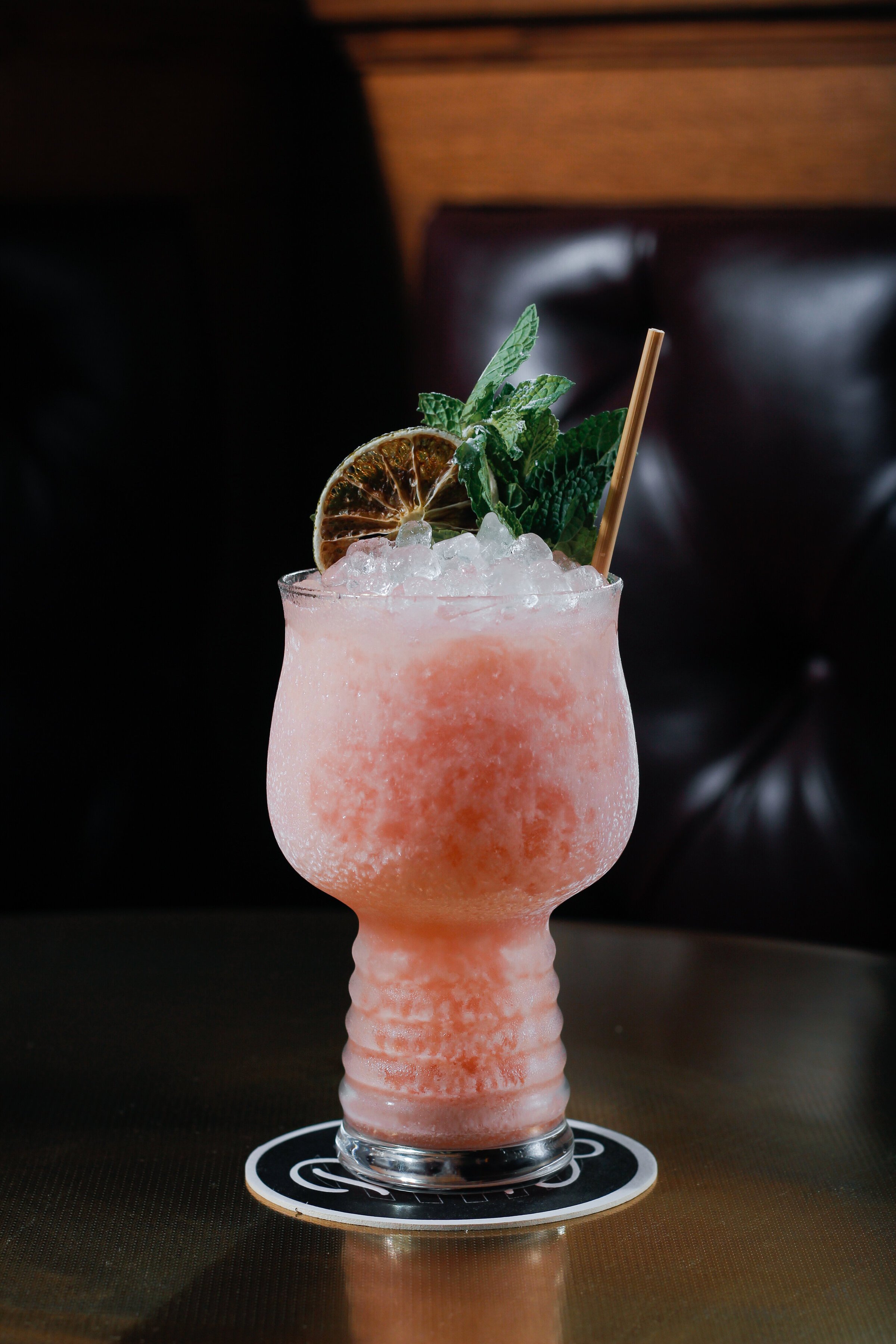 A pink slushy cocktail garnished with a slice of dried lime, fresh mint leaves, and a wooden straw, served in a glass with a textured design, on a dark coaster.