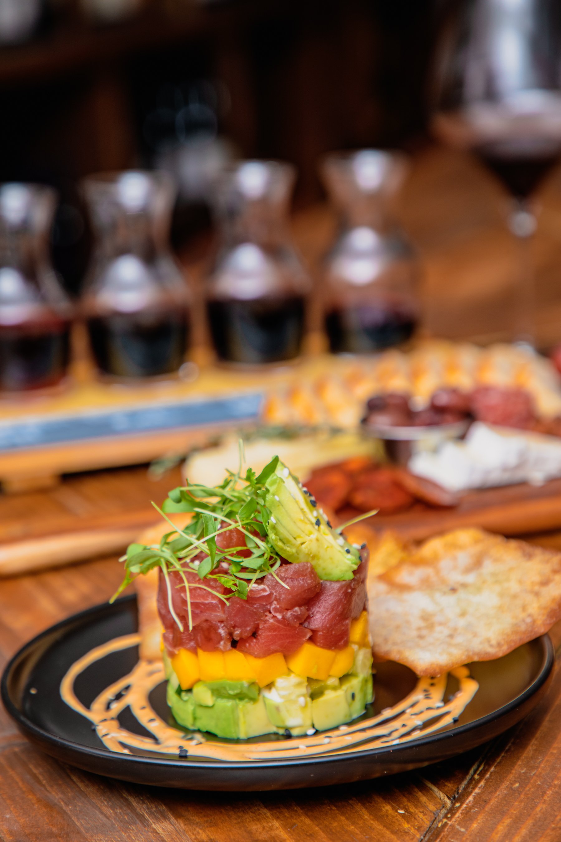 A plate of tuna tartare with sliced avocado, mango, and microgreens garnished with a slice of jalapeño, served with a crispy flatbread on the side. In the background, there are small glass bottles of red wine and a variety of other appetizers on a wo